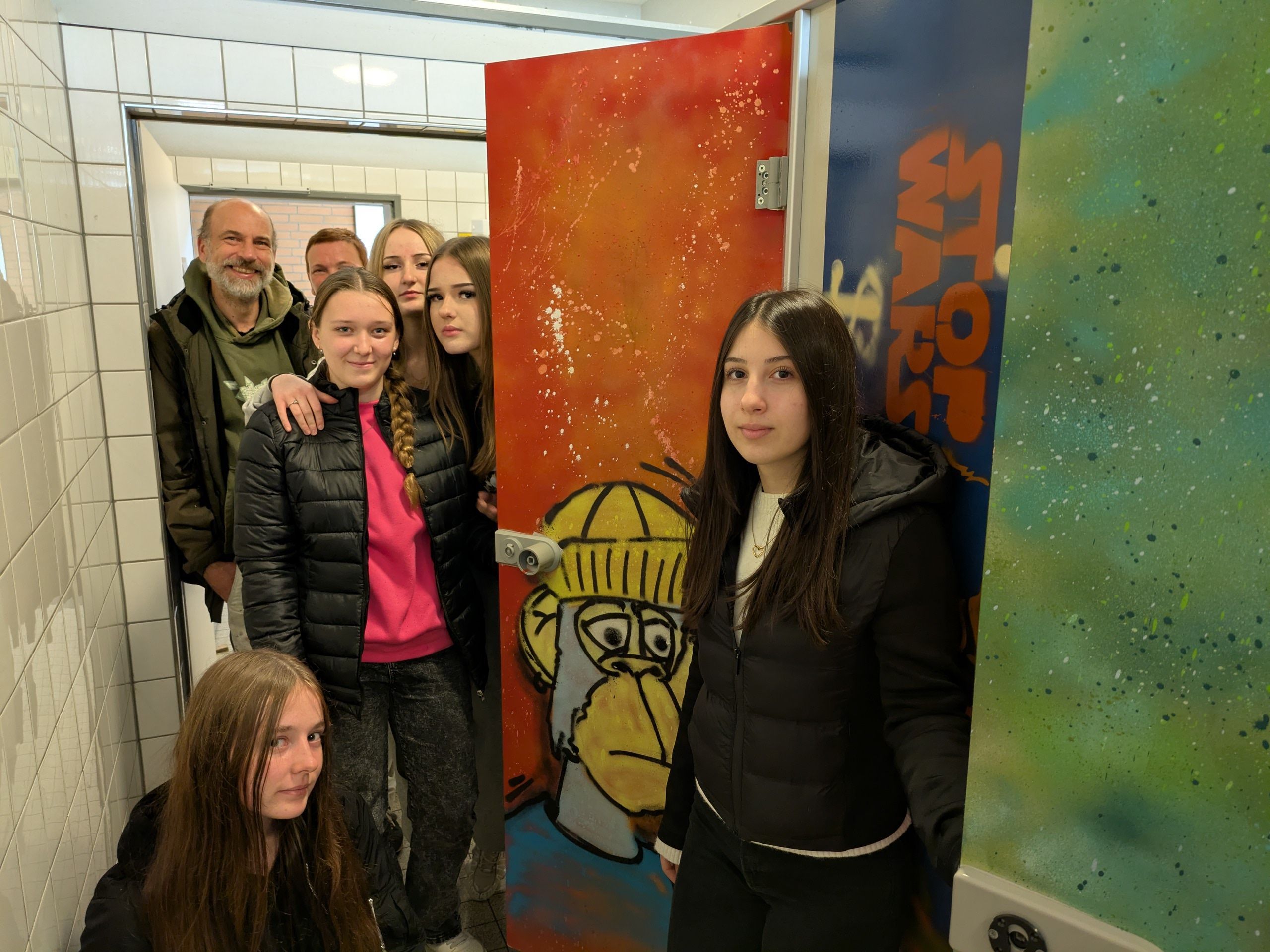 Schülerinnen und ein Mann stehen in einer Schultoilette mit Graffiti.
