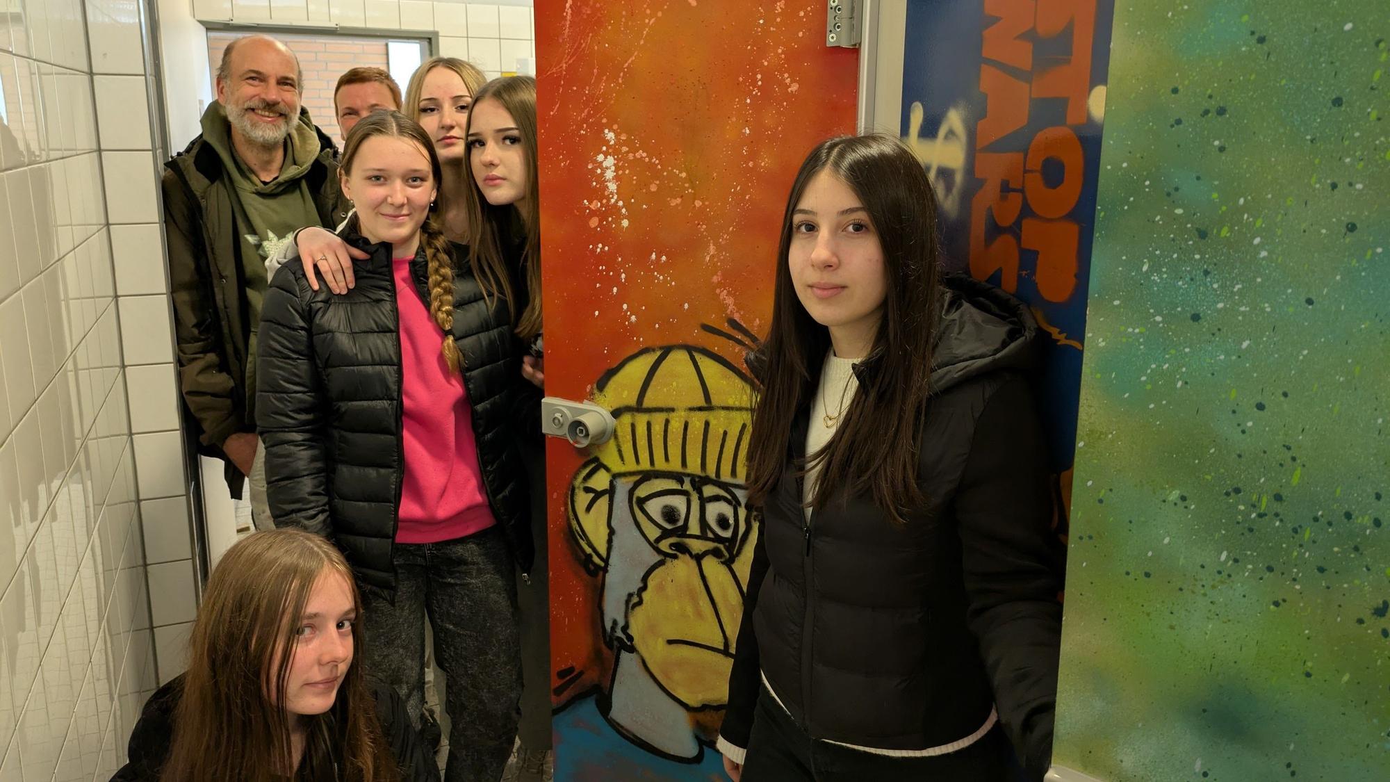 Schülerinnen und ein Mann stehen in einer Schultoilette mit Graffiti.