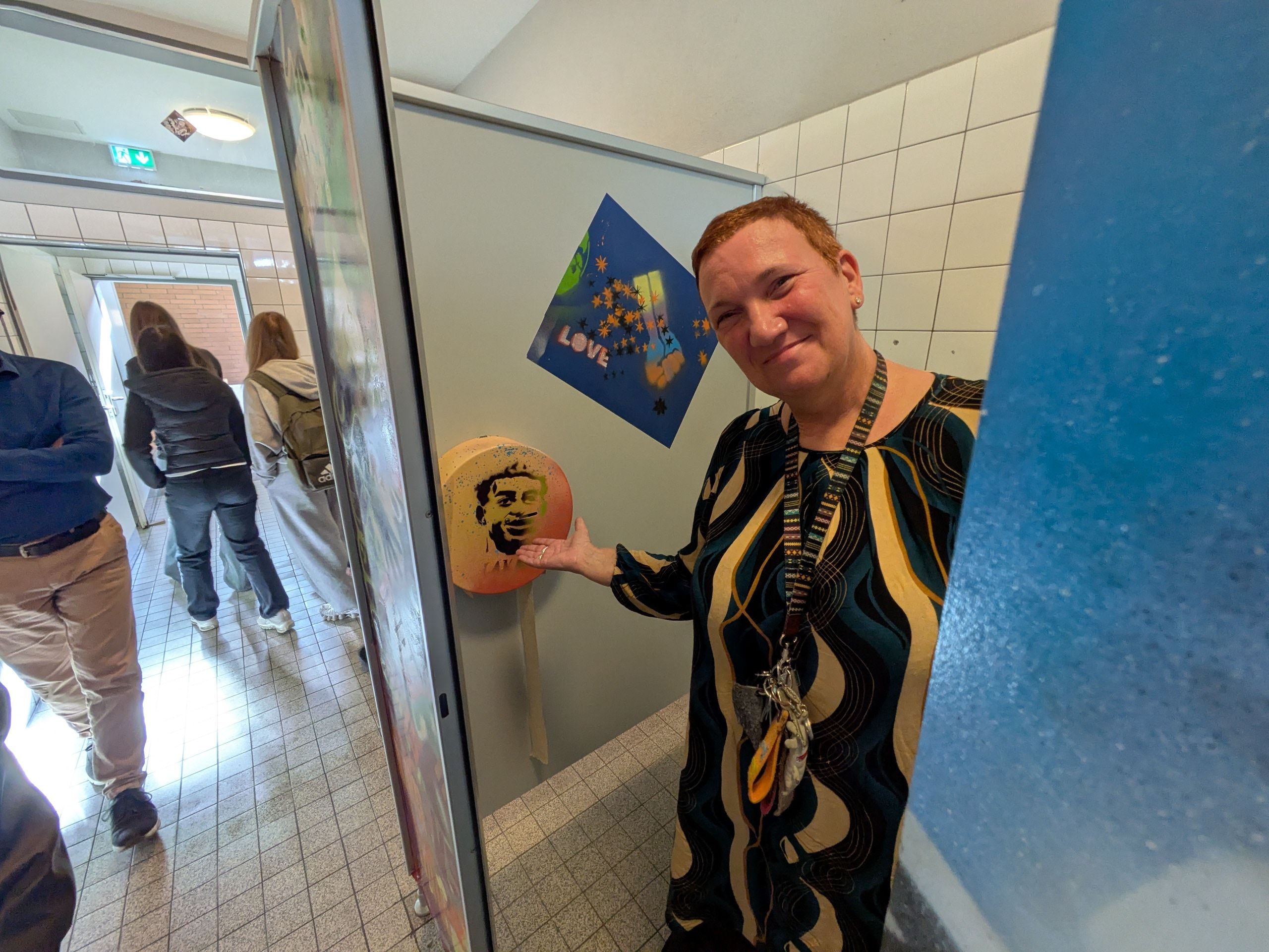 Eine Frau steht in einer Toilette und zeigt auf einen gestalteten Klopapierhalter.
