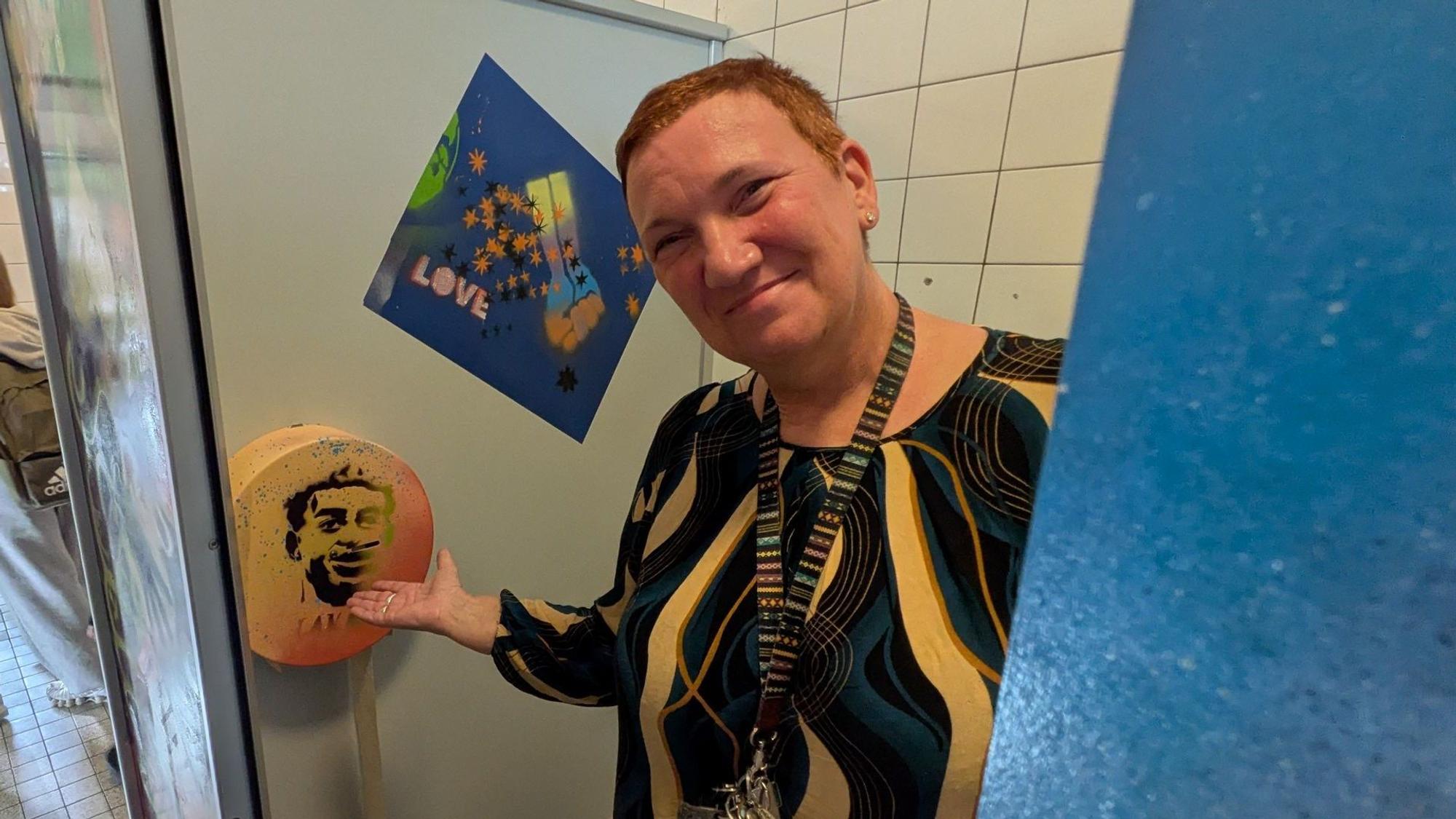 Eine Frau steht in einer Toilette und zeigt auf einen gestalteten Klopapierhalter.