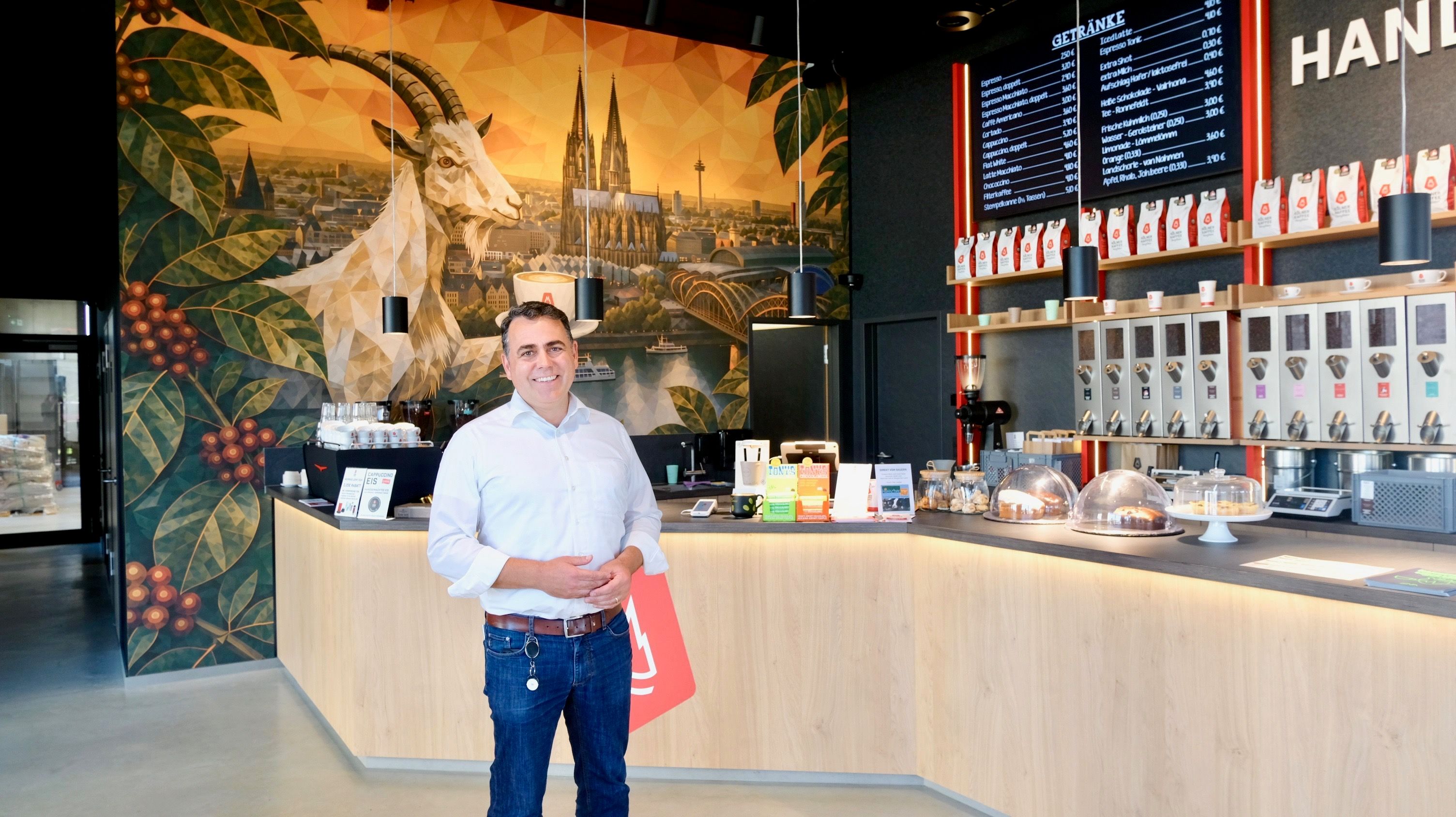 Ein Mann im weißen Hemd steht vor einem großen Verkaufstresen, hinter ihm ziert eine große Fototapete mit Dom und Geißbock die Wand.