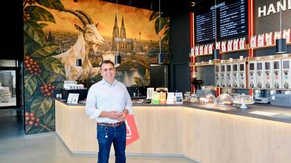 Ein Mann im weißen Hemd steht vor einem großen Verkaufstresen, hinter ihm ziert eine große Fototapete mit Dom und Geißbock die Wand