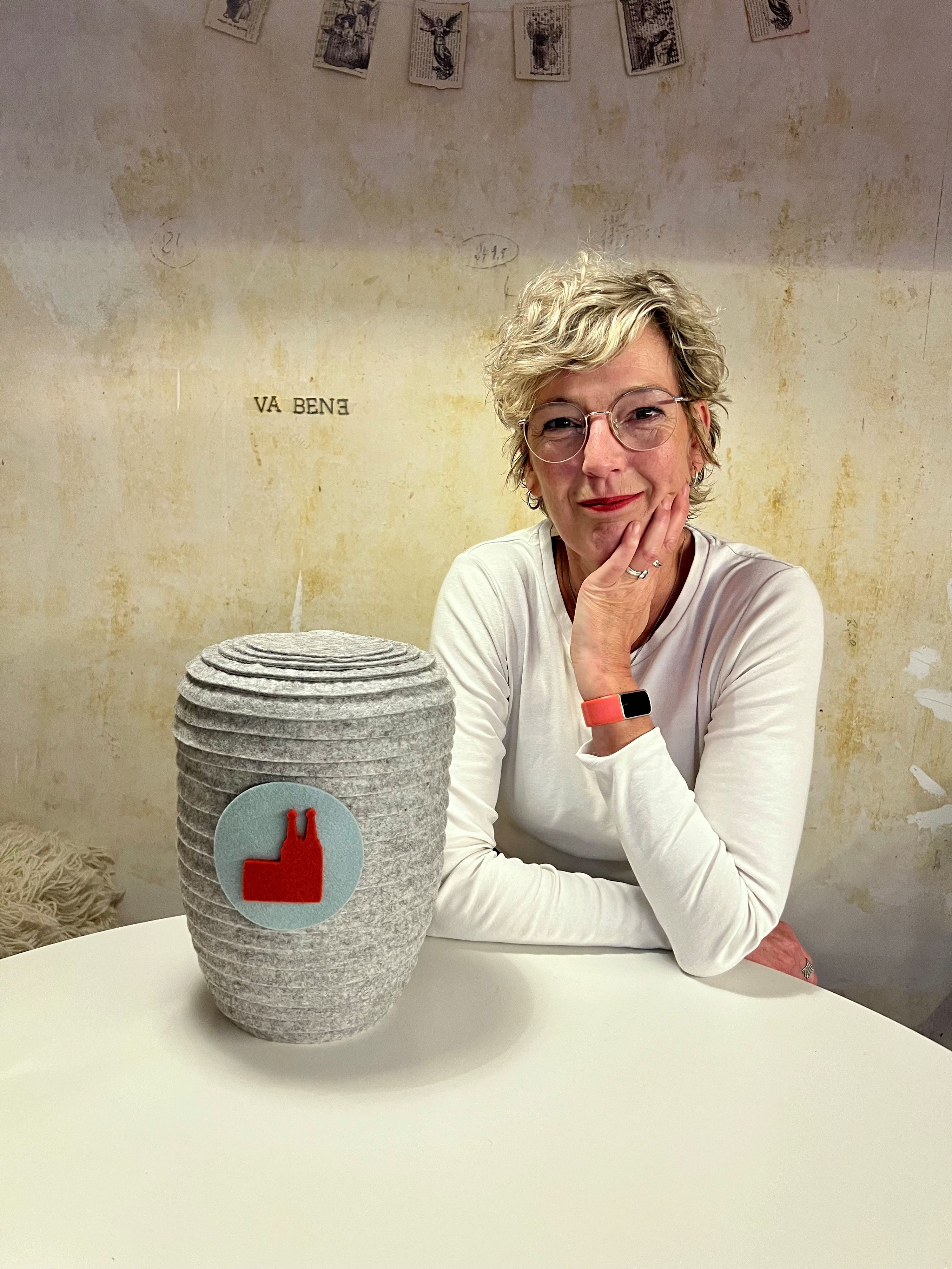 Eine Frau im weißen T-Shirt sitzt an einem Tisch, vor ihr steht eine Urne im Filzmantel.