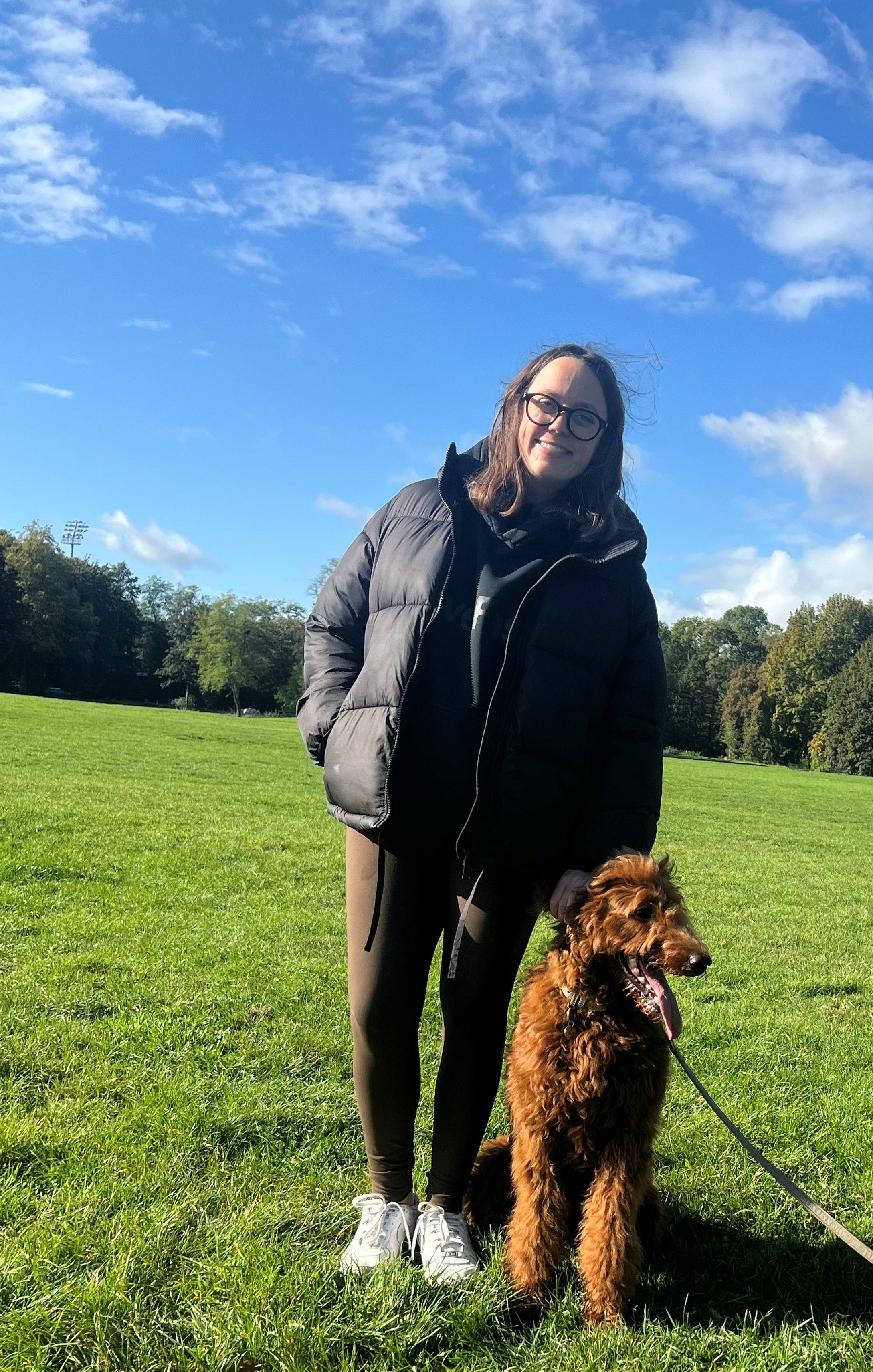 Eien Frau mit Steppjacke steht auf einer Wiese, neben sich ein großer brauner Pudel.