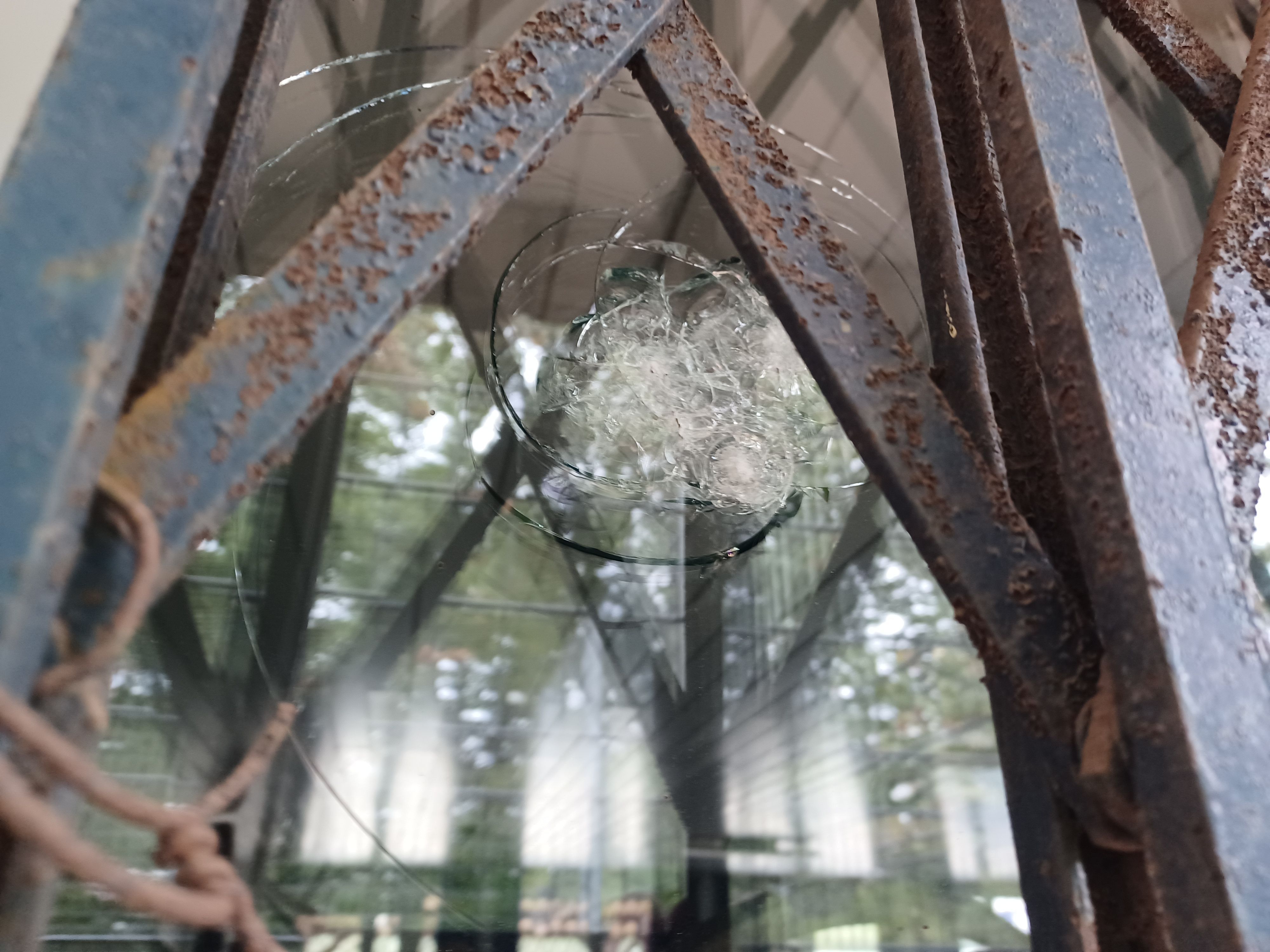 Zu sehen ist eine Glasscheibe mit einem großen Einschlagloch und radialen Sprüngen, eingefasst von verrosteten Metallstreben.