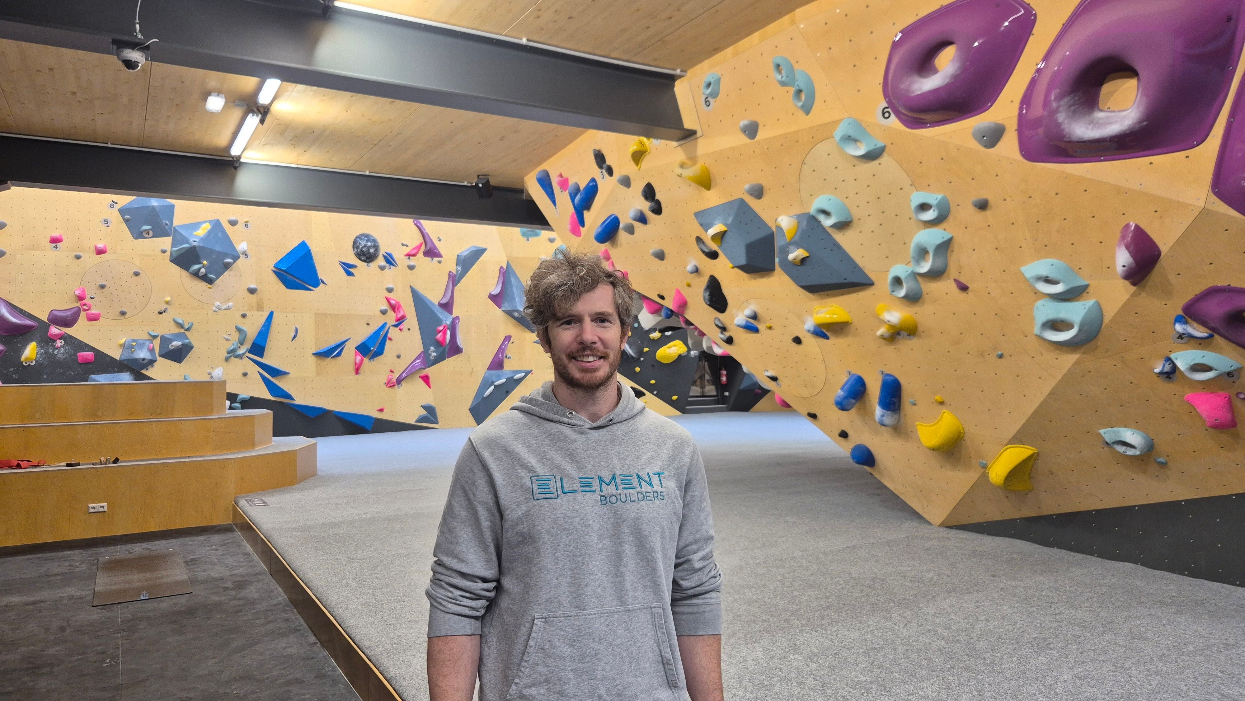 Ein lächelnder Mann mit grauem Kapuzenpullover („Element Boulders“) steht in einer Boulderhalle vor einer Kletterwand mit bunten Griffen.