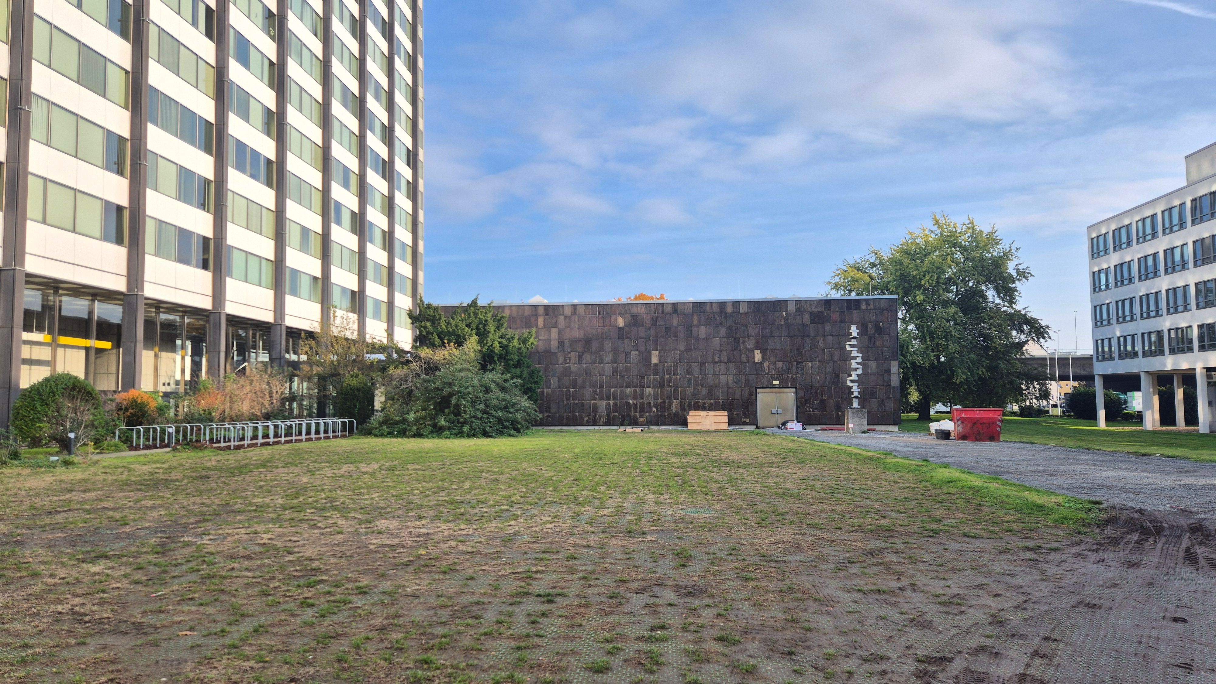 Eine grasbewachsene Freifläche zwischen einem hohen Bürogebäude links, davor eine mit braunem Quarzit verkleidete Halle.