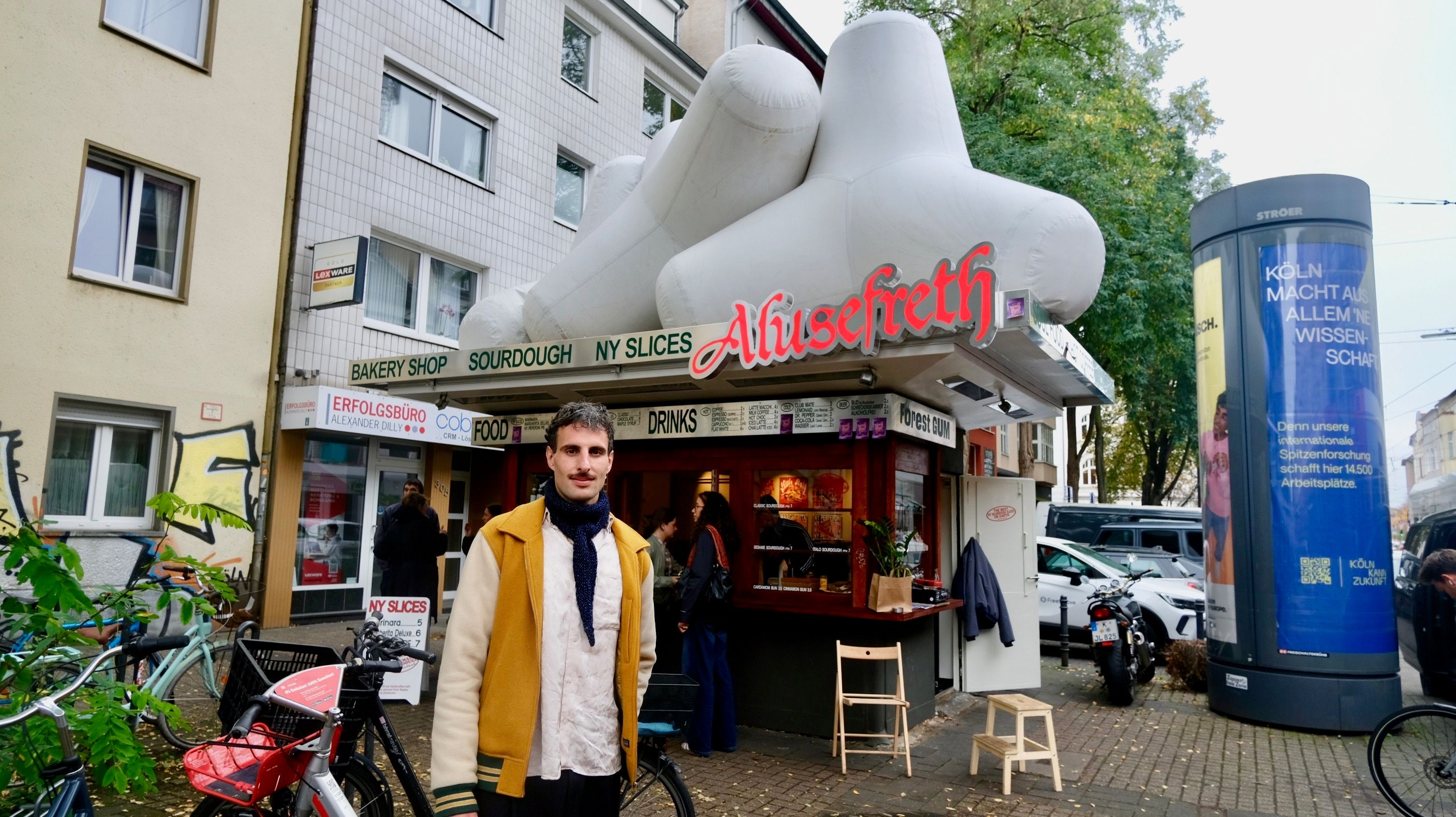 Ein junger Mann mit dunklem Haar und Schnurrbart steht vor einem Kiosk mit dreieckigen Figuren auf dem Dach.