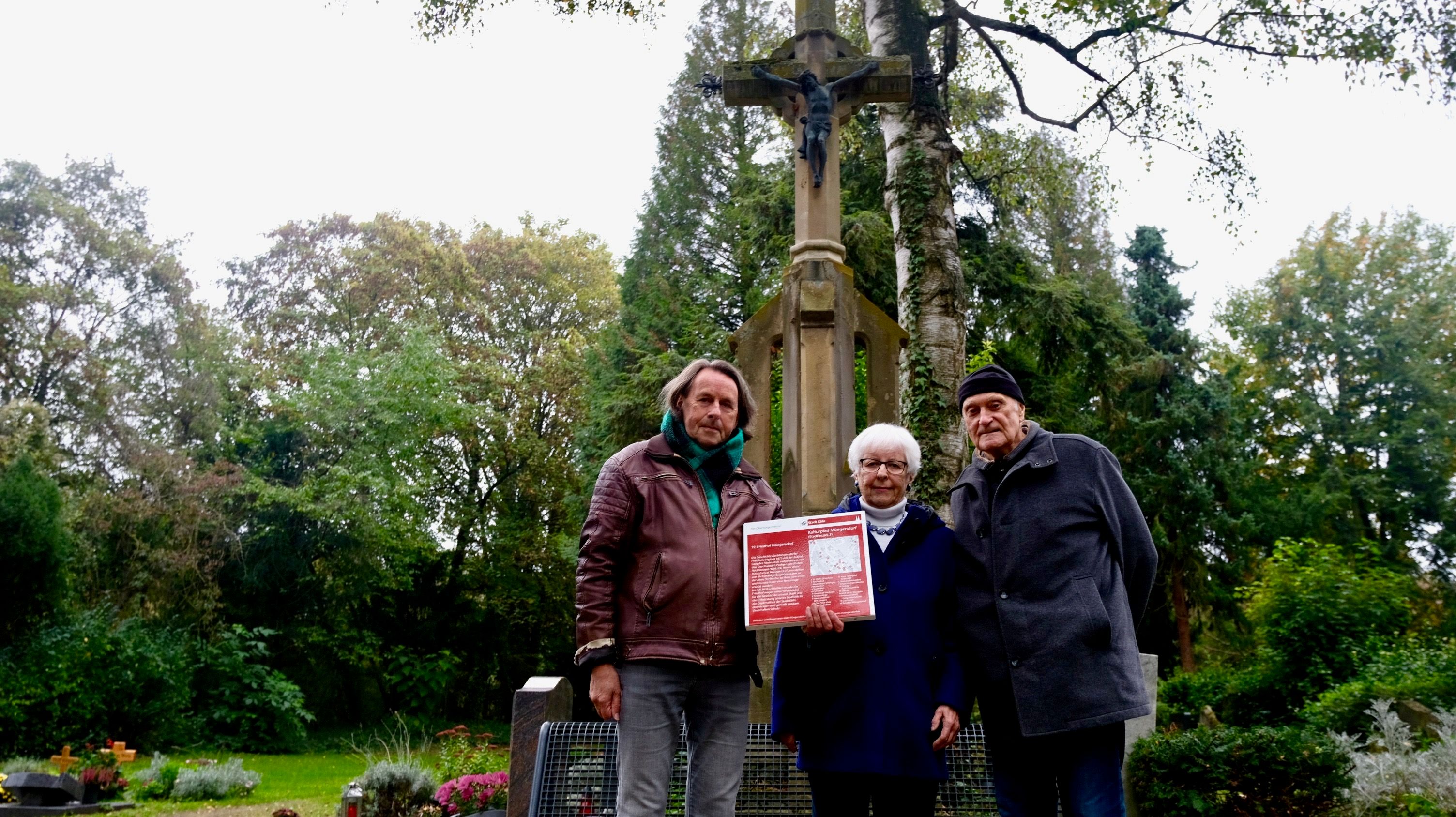 Zwei Männer und eine Frau zeigen eine rote Infotafel, die den Müngersdorfer Friedhof als Denkmal ausweist.