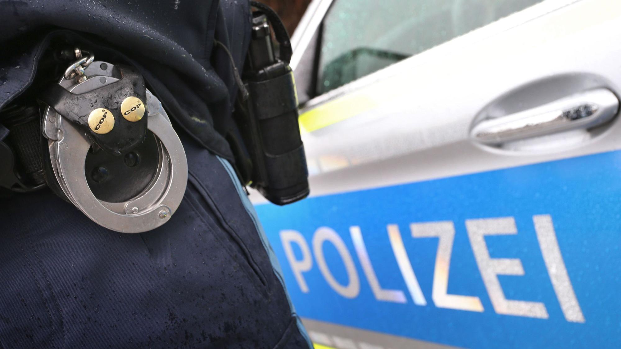 Ein Polizist mit Handschellen und einer Pistole am Gürtel steht vor einem Streifenwagen.