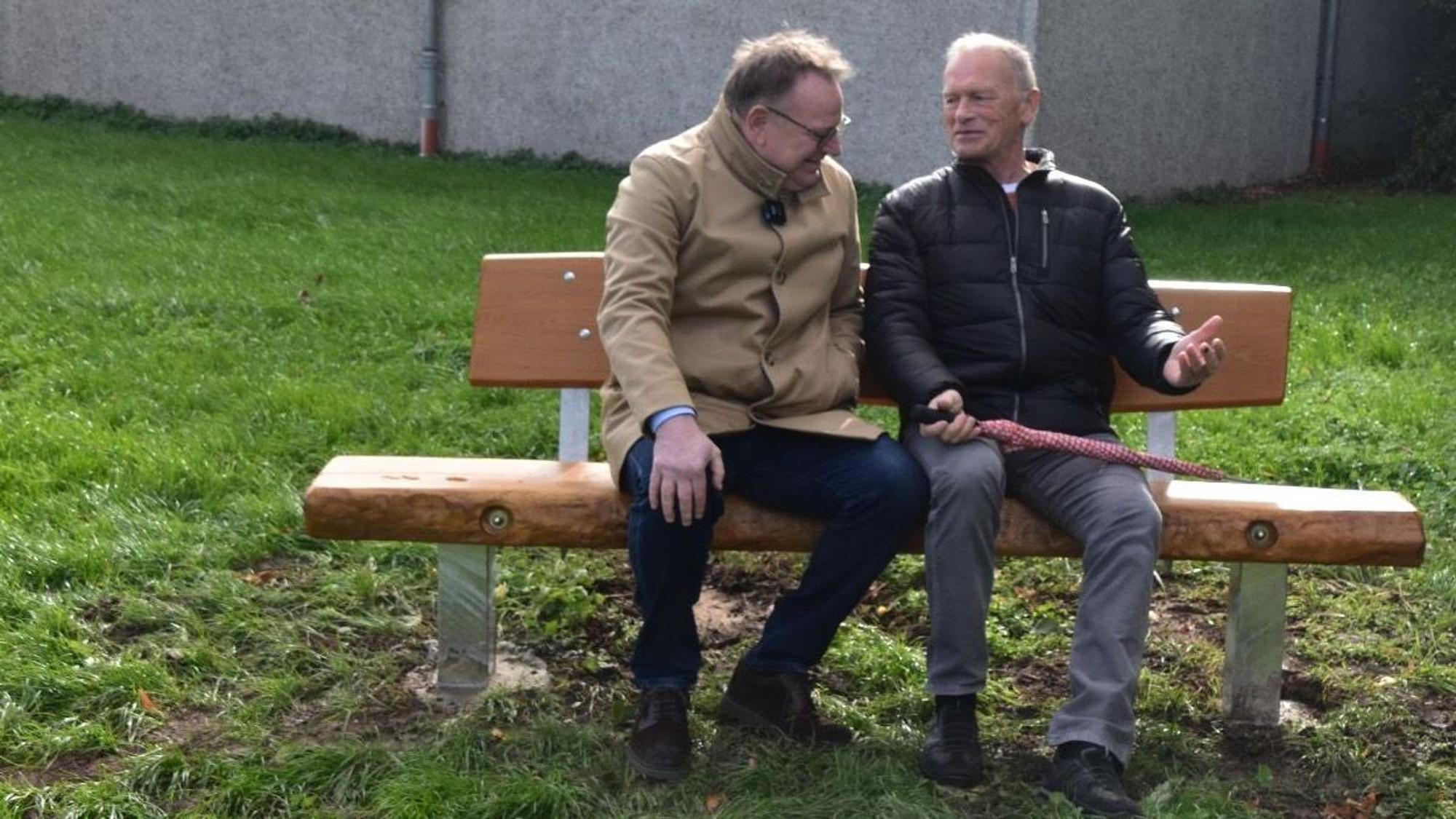 Zwei Männer sitzen nebeneinander auf einer Bank. Ein Kameramann hält ein Mikrofon in ihre Richtung.