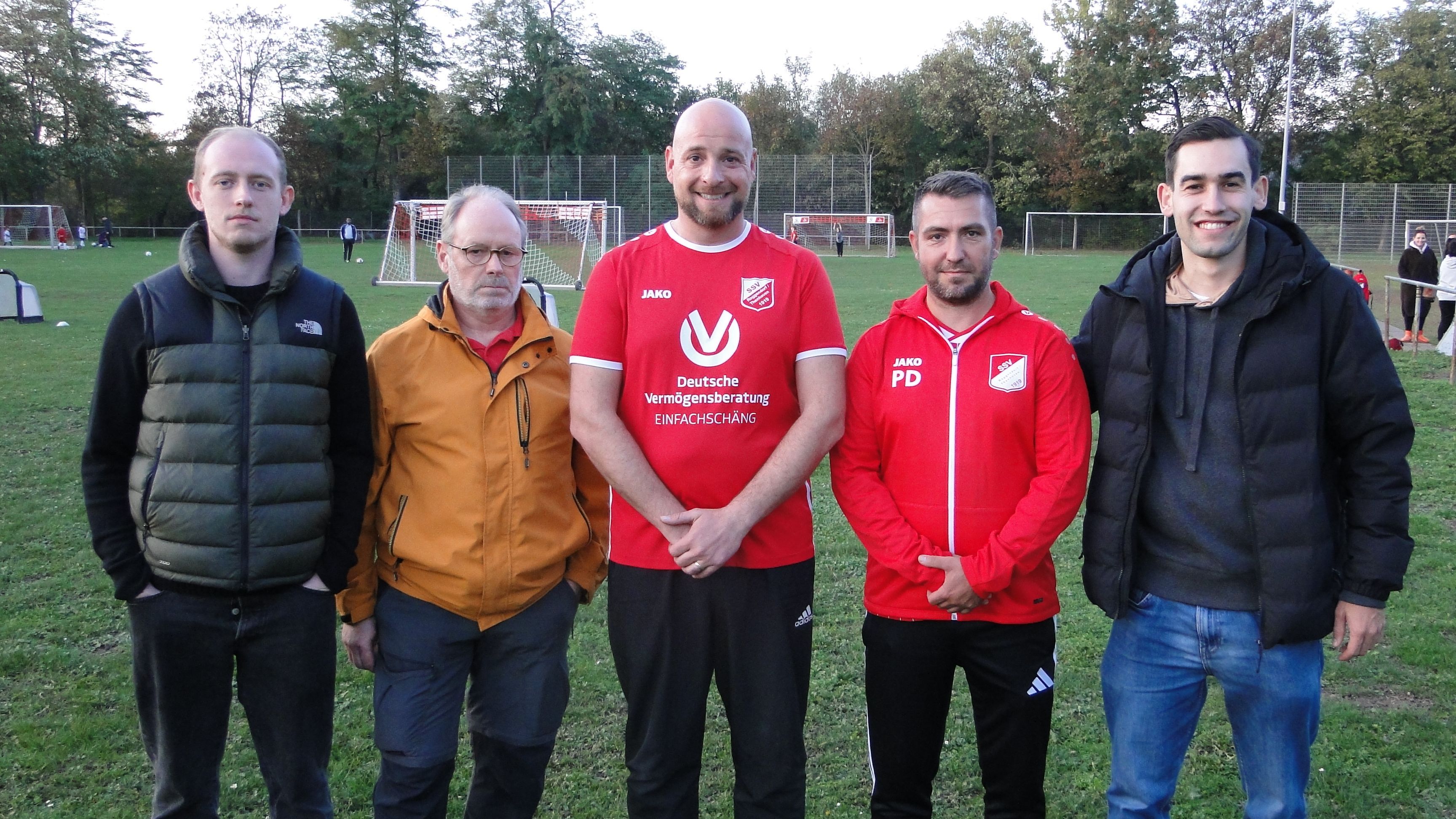 Fünf Männer stehen nebeneinander auf einem Naturrasenplatz, zwei davon in roten Trikots bzw. Trainingsjacken mit Vereinslogo. Im Hintergrund spielen weitere Personen und Tore sind zu sehen.