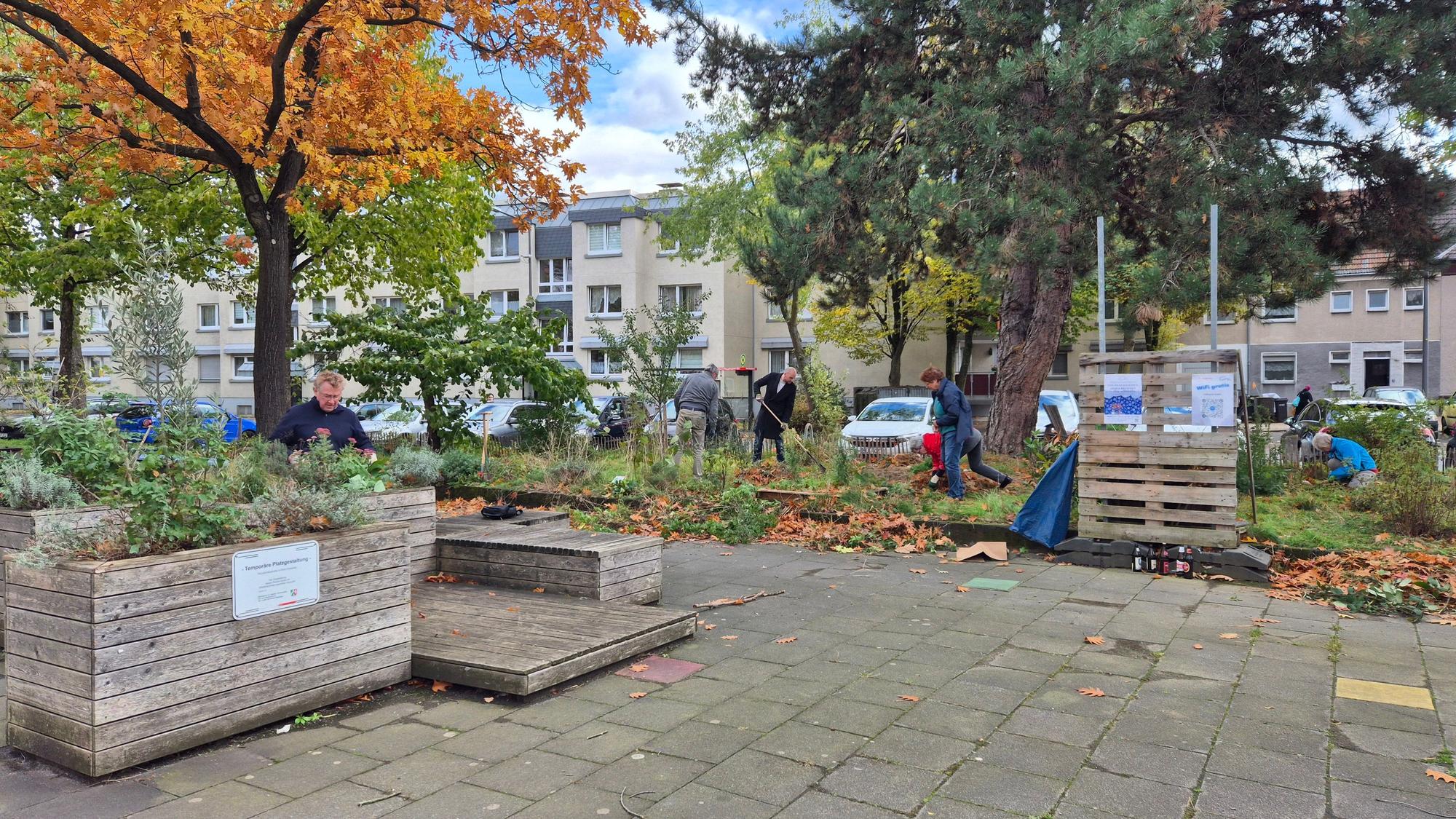 Mehrere Menschen arbeiten im Herbst auf einem begrünten Platz mit Hochbeeten, Holzpaletten und Laub. Im Hintergrund stehen Wohnblöcke.