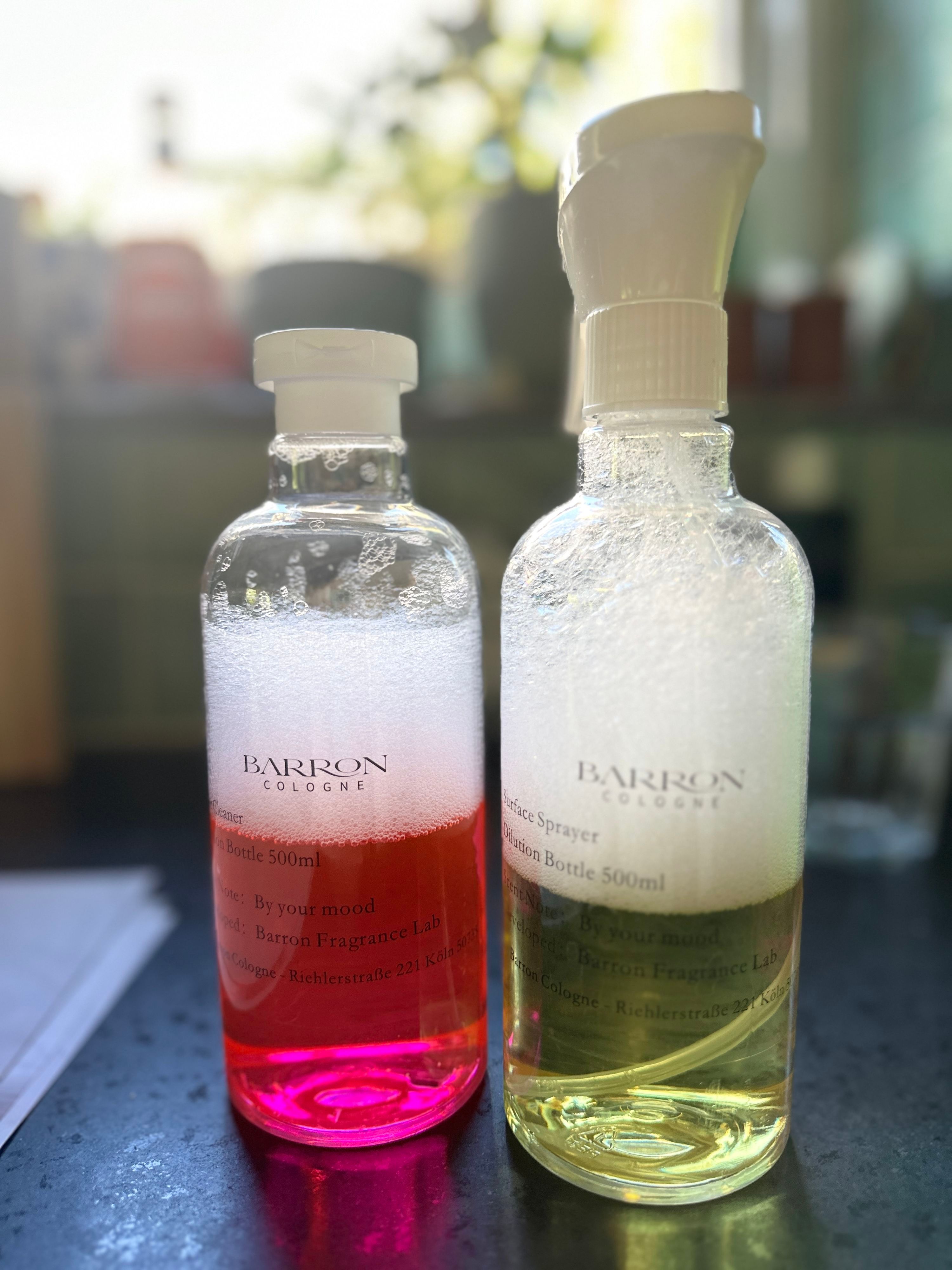 Zwei Flaschen mit Barron-Cologne-Etikett stehen nebeneinander auf einer dunklen Arbeitsfläche: links eine klare Flasche mit pinkfarbener Flüssigkeit und Schaum, rechts eine Sprühflasche mit gelblicher Flüssigkeit und Schaum.