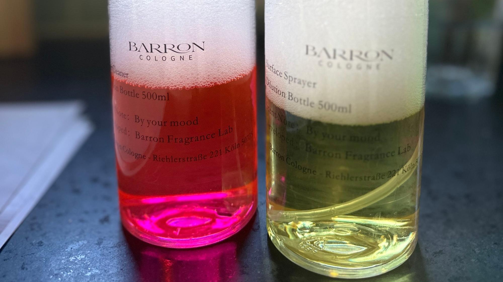 Zwei Flaschen mit Barron-Cologne-Etikett stehen nebeneinander auf einer dunklen Arbeitsfläche: links eine klare Flasche mit pinkfarbener Flüssigkeit und Schaum, rechts eine Sprühflasche mit gelblicher Flüssigkeit und Schaum.