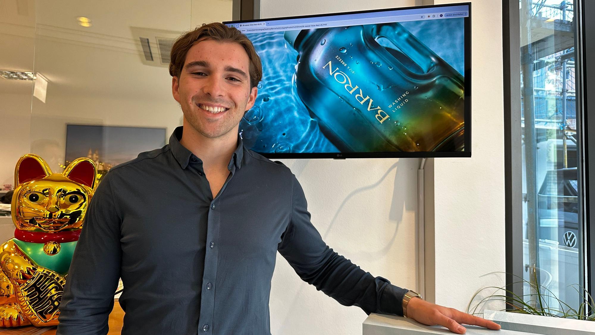 Ein lächelnder junger Mann im dunkelgrauen Hemd steht in einem hellen Raum neben eeinergoldenen Winkekatzen-Figur und einem Monitor mit der Werbung für „Barron Washing Liquid“.
