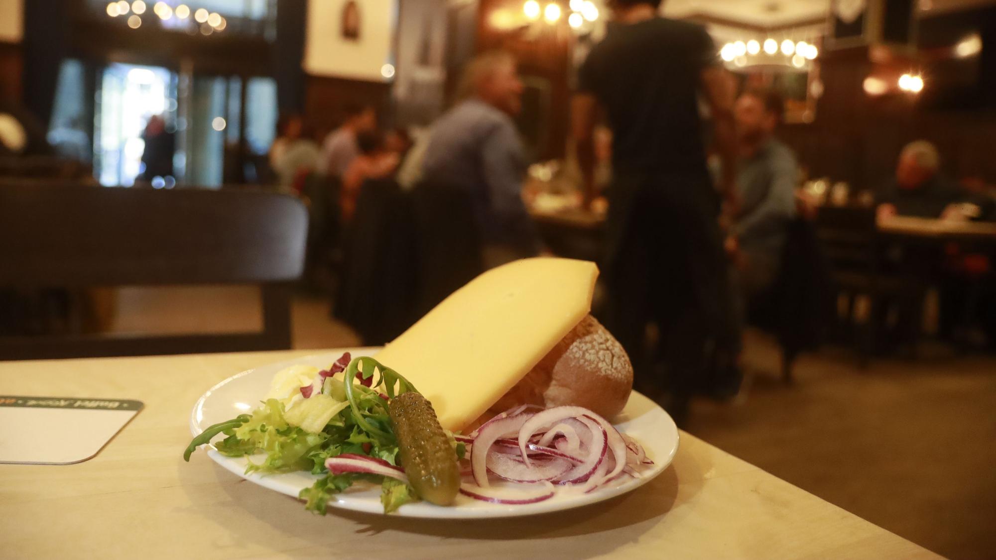 Auf einem Teller liegen ein Roggenbrötchen, eine dicke Scheibe mittelalter Gouda, Zwiebeln sowie eine Gewürzgurke.