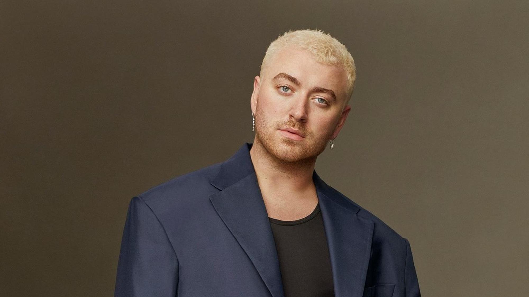 Sam Smith mit blau/schwarzem Anzug vor einer braunen Wand. Er hat blondierte Haare und schaut ernst in die Kamera.