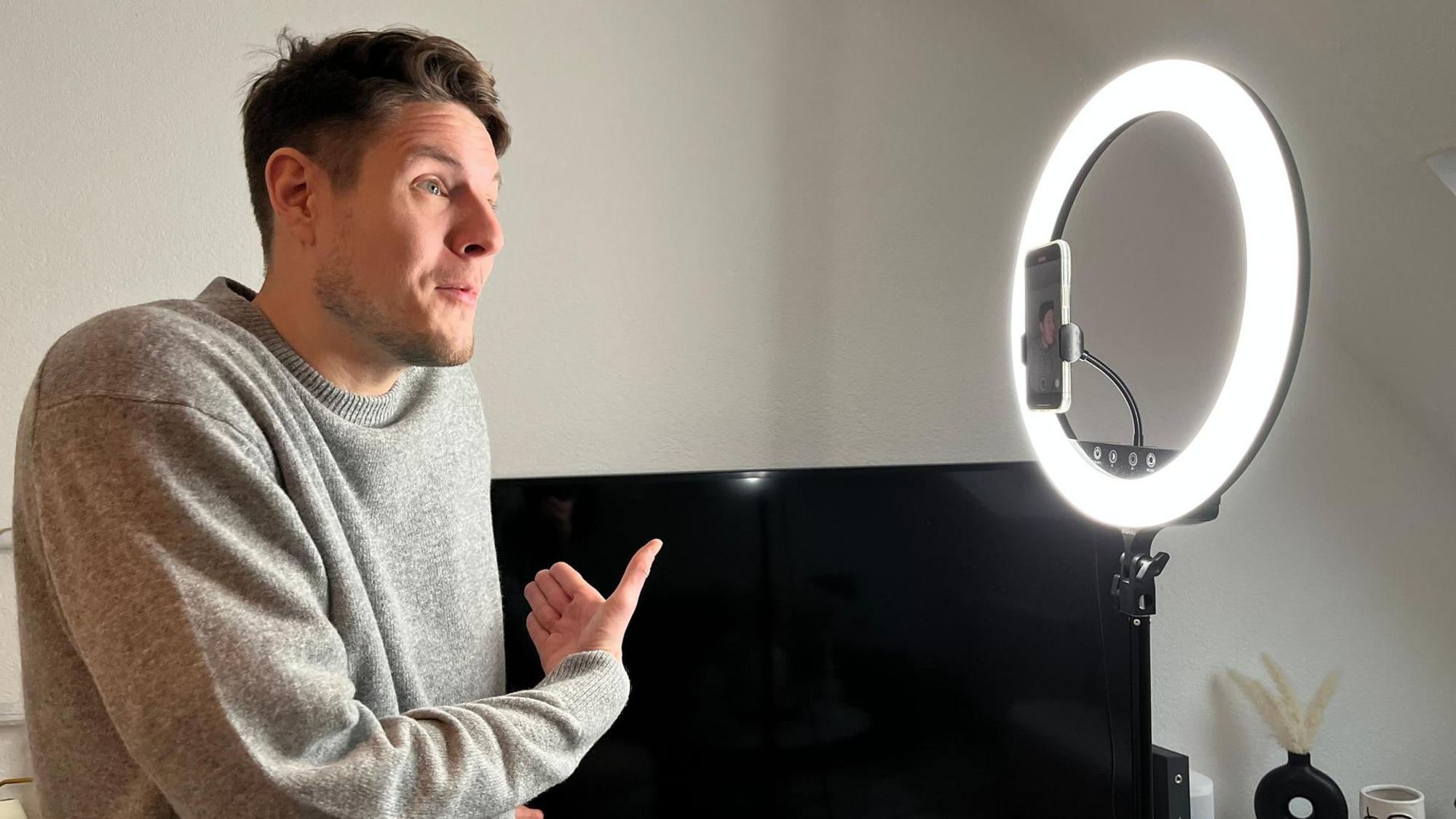 Ein Mann im grauen Pulli, mit braunen Haaren und Dreitagebart steht vor einem Ringlicht und spricht in die Handykamera.