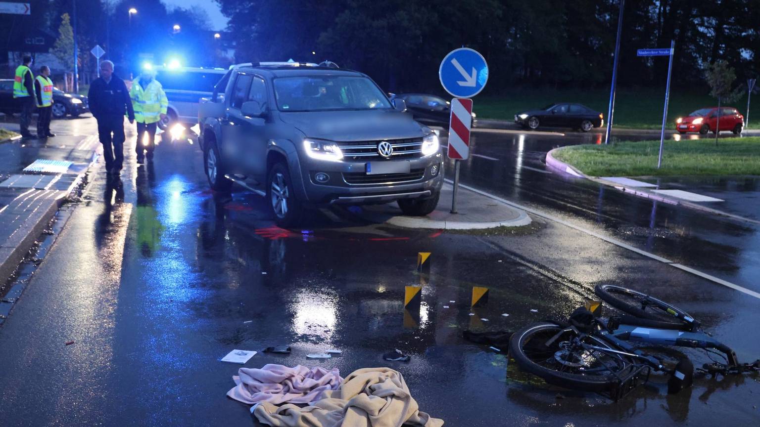Im Dunkeln steht ein Auto halb auf einer Verkehrsinsel, ein Pedelec-Rad liegt auf dem Boden, Decken daneben. Im Hintergrund leuchtdn die Polizeisirenen blau.