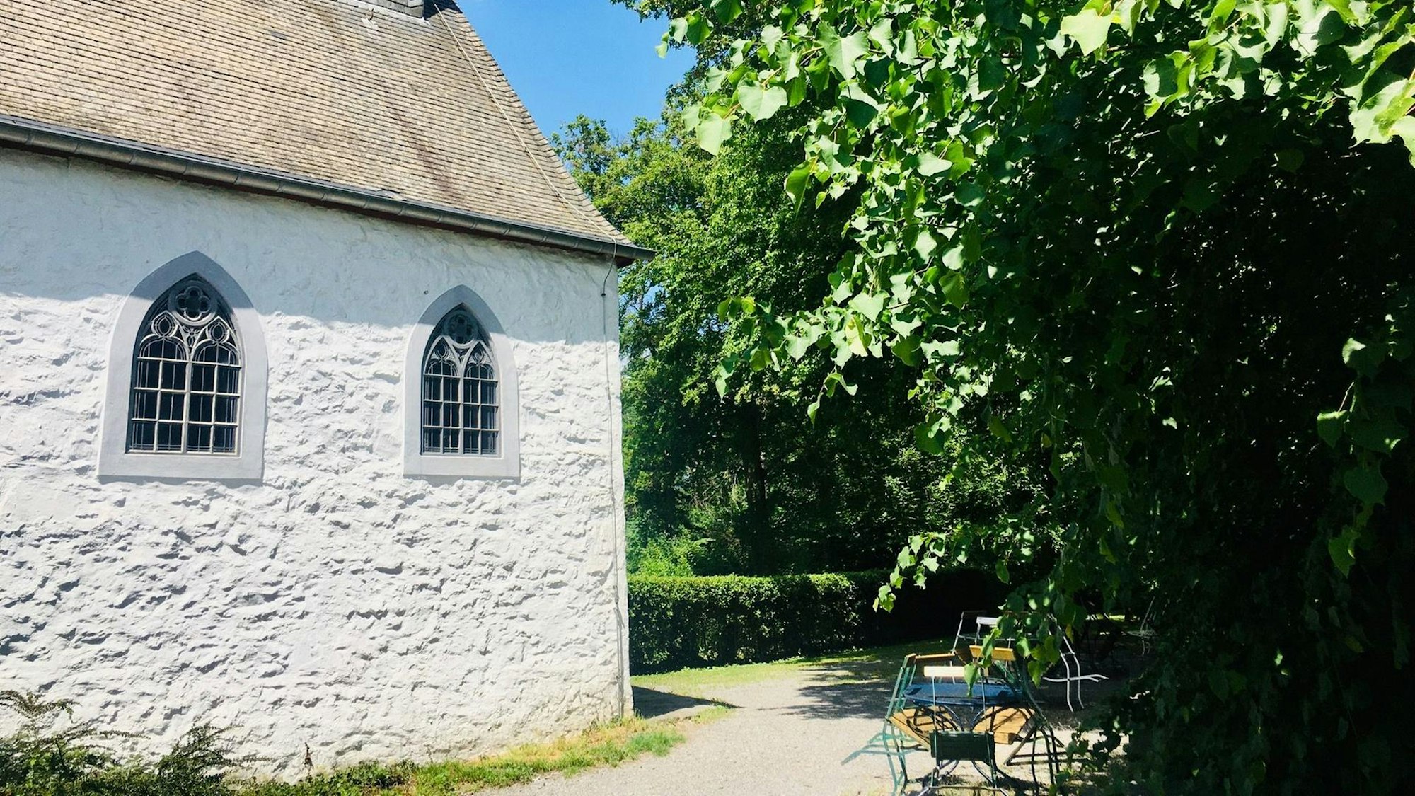 Eine Ecke der Kapelle, daneben ein Weg mit Stühlen am Rand und Bäume