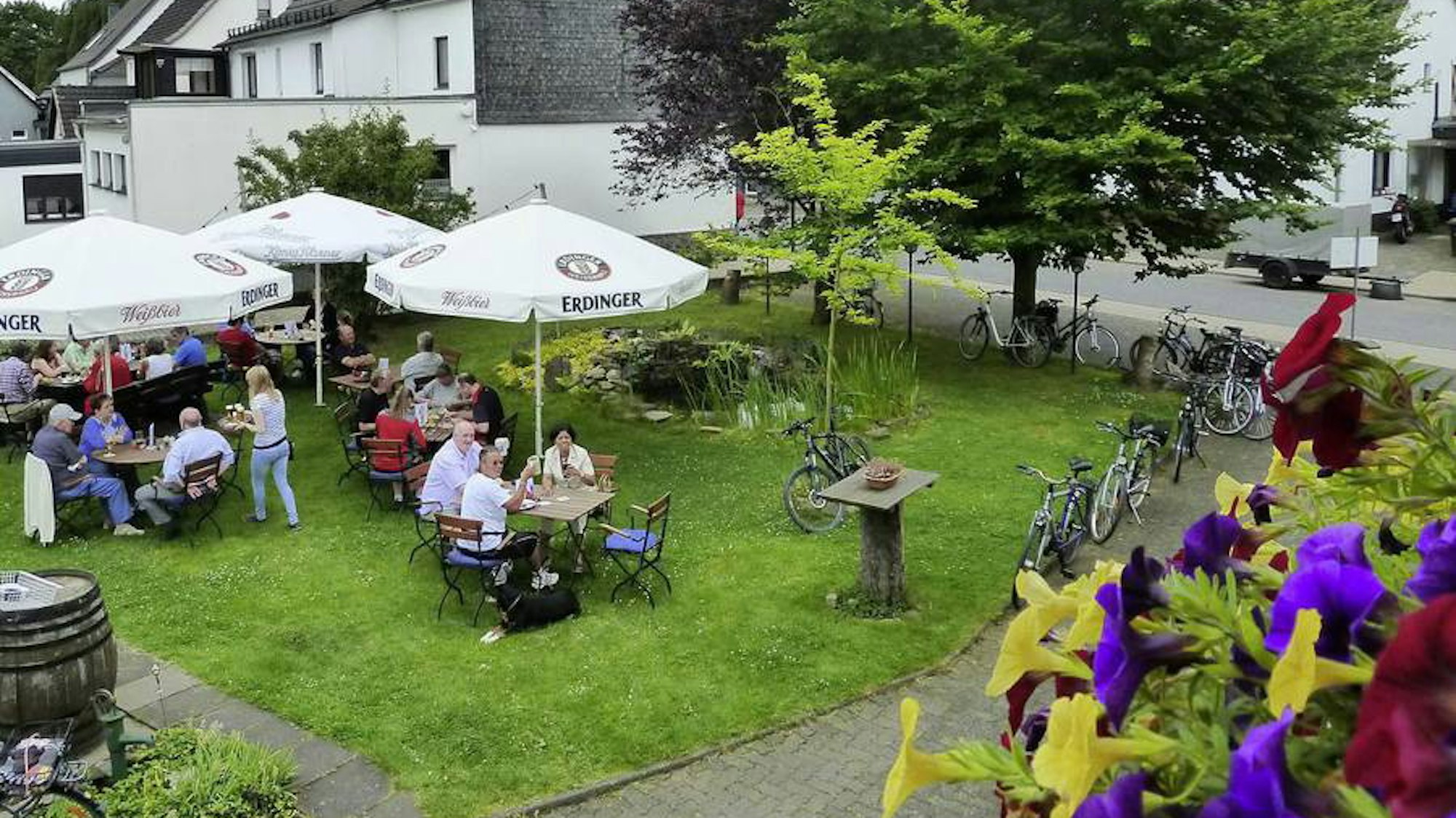 Auf einer Wiese stehen Tische und Stühle unter Sonnenschirmen. Viele Menschen sitzen dort.