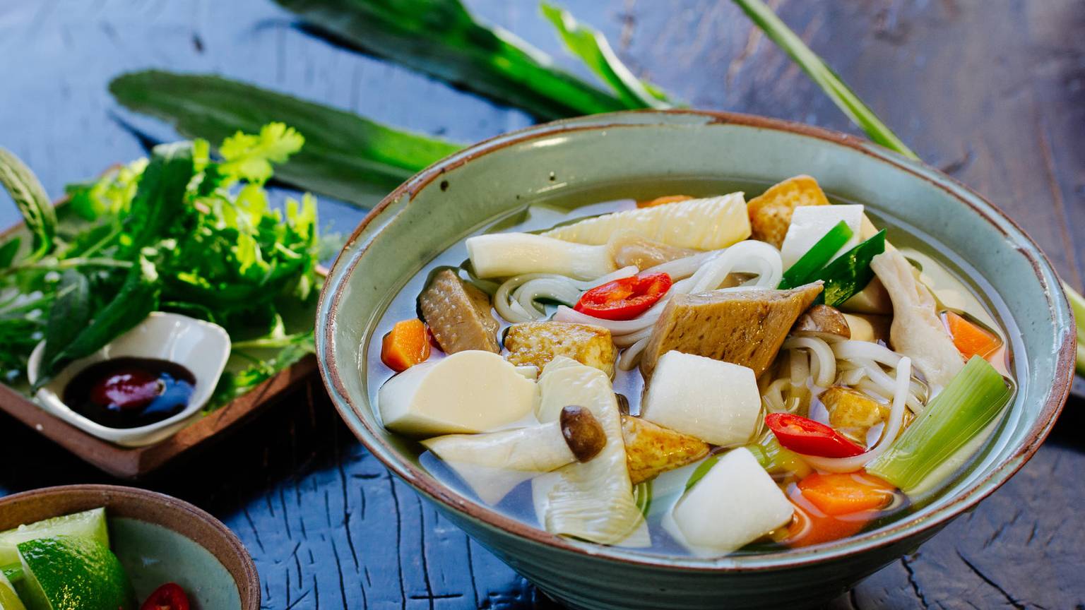 Eine Pho-Suppe mit vegetarischen Zutaten steht auf einem Tisch. Daneben stehen Kräuter und Soßen.