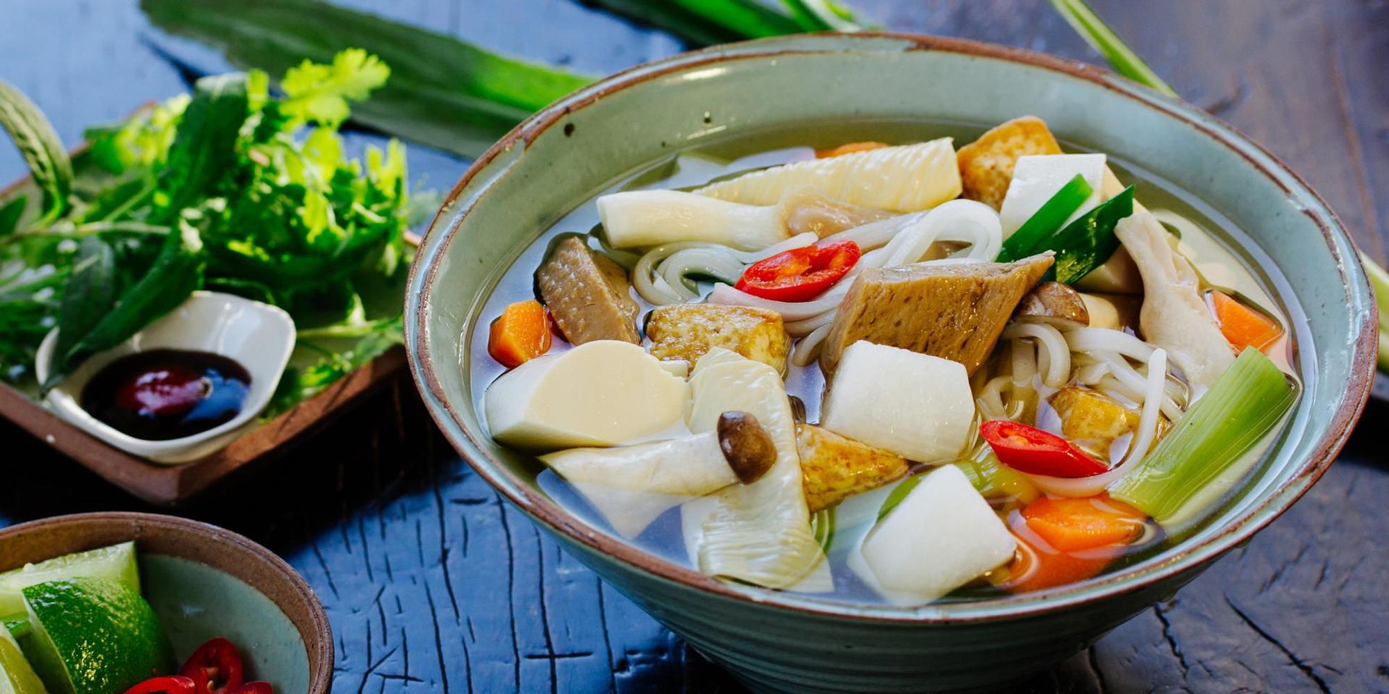 Eine Pho-Suppe mit vegetarischen Zutaten steht auf einem Tisch. Daneben stehen Kräuter und Soßen.