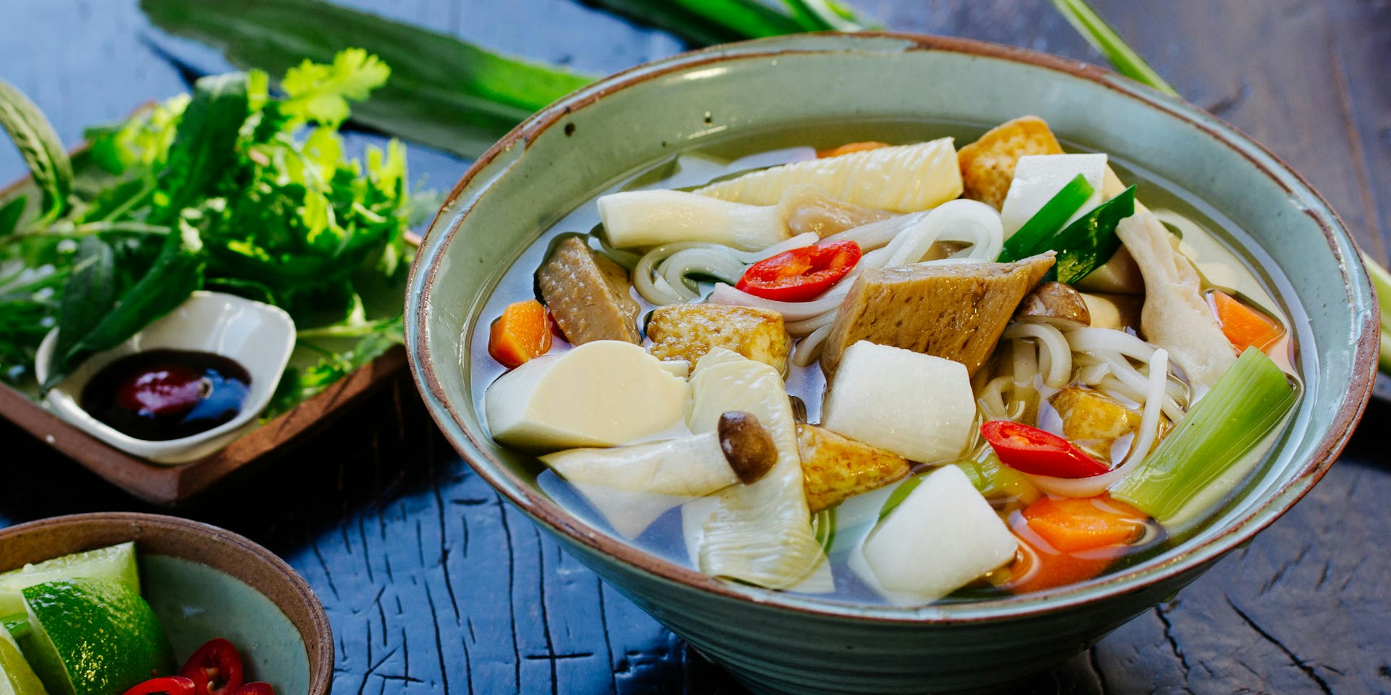 Eine Pho-Suppe mit vegetarischen Zutaten steht auf einem Tisch. Daneben stehen Kräuter und Soßen.