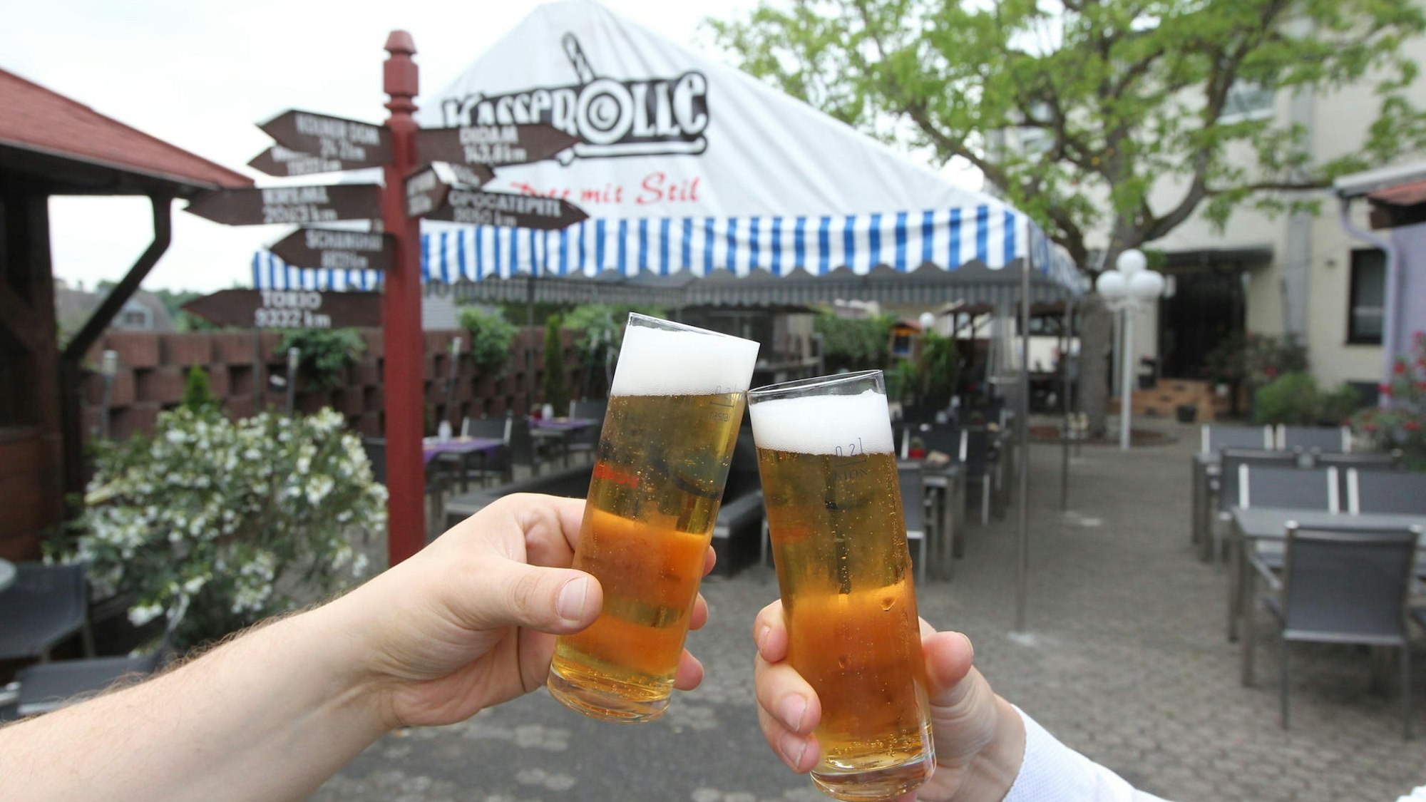 Zwei Hände halten jeweils ein Kölschglas mit Bier in der Hand. Im Hintergrund stehen auf einer Terrasse Tische, Stühle und ein Zelt.