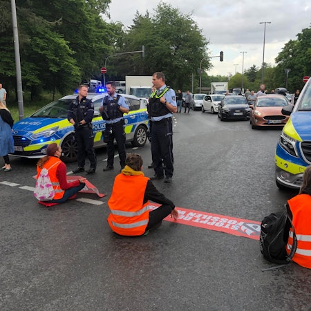 Vier Klima-Aktivisten der Letzten Generation sitzen in orangefarbenen Warnwesten auf der Inneren Kanalstraße und blockieren die Fahrbahn.