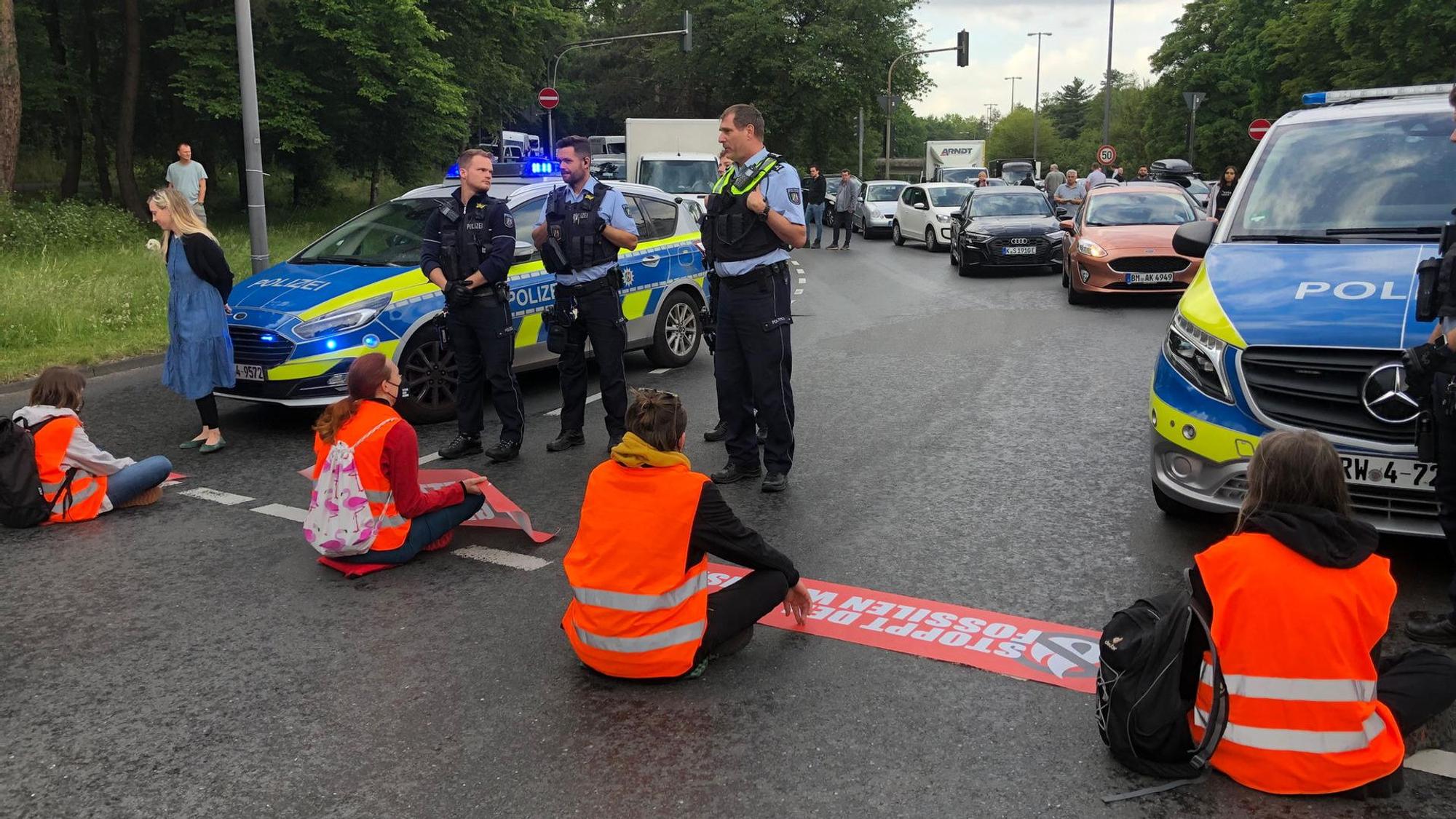 Vier Klima-Aktivisten der Letzten Generation sitzen in orangefarbenen Warnwesten auf der Inneren Kanalstraße und blockieren die Fahrbahn.