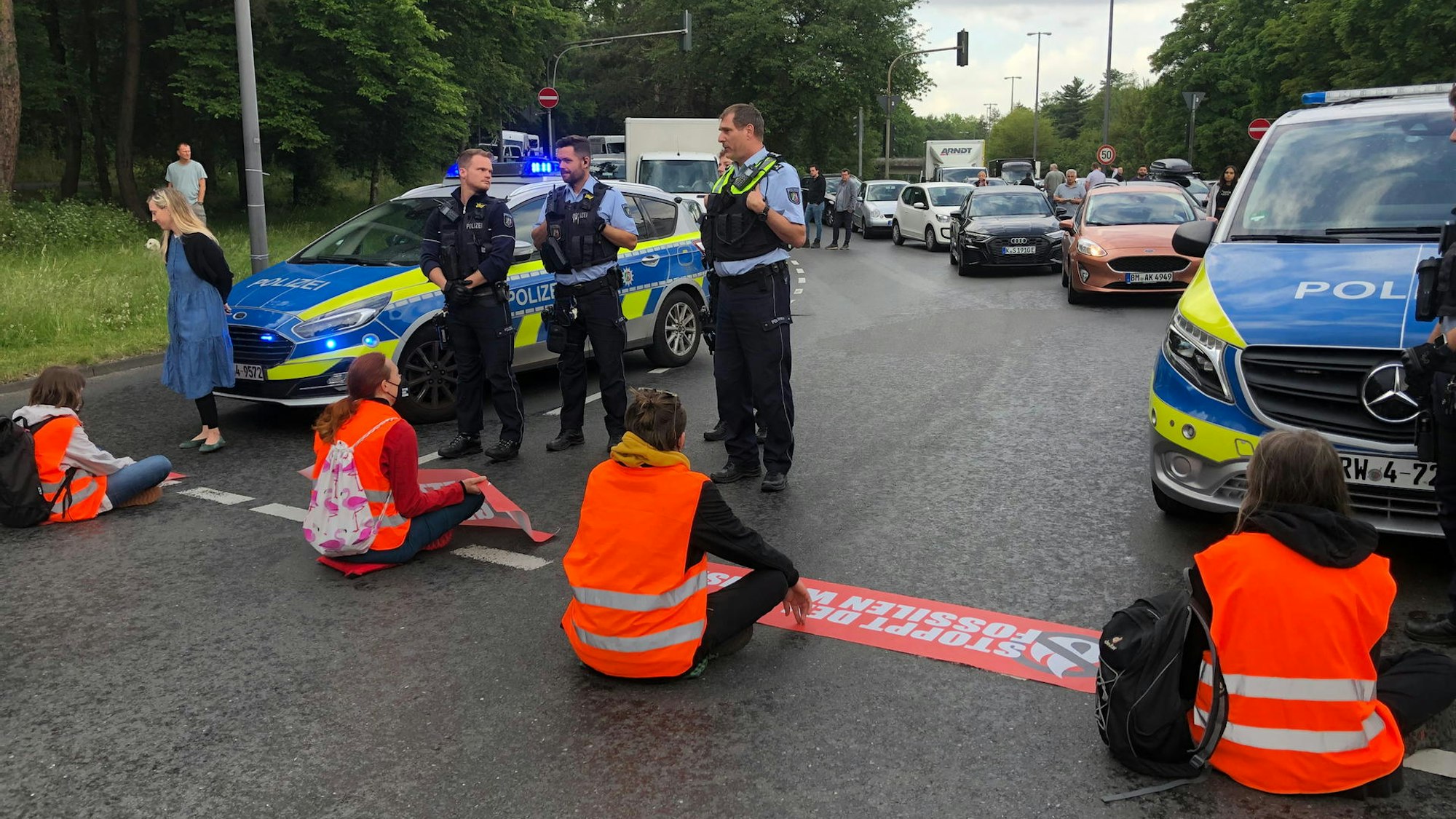 Vier Klima-Aktivisten der Letzten Generation sitzen in orangefarbenen Warnwesten auf der Inneren Kanalstraße und blockieren die Fahrbahn.