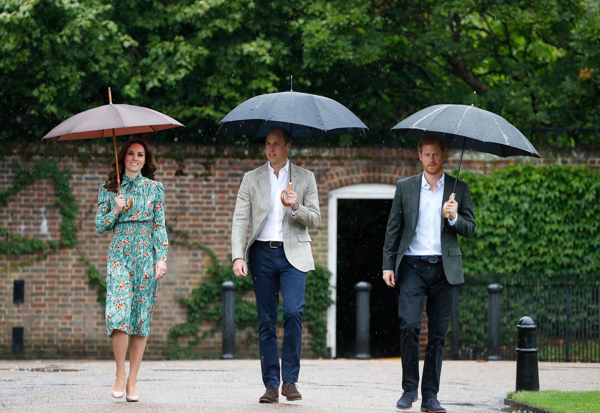William, Harry und Kate gehen nebeneinander mit Schirmen.