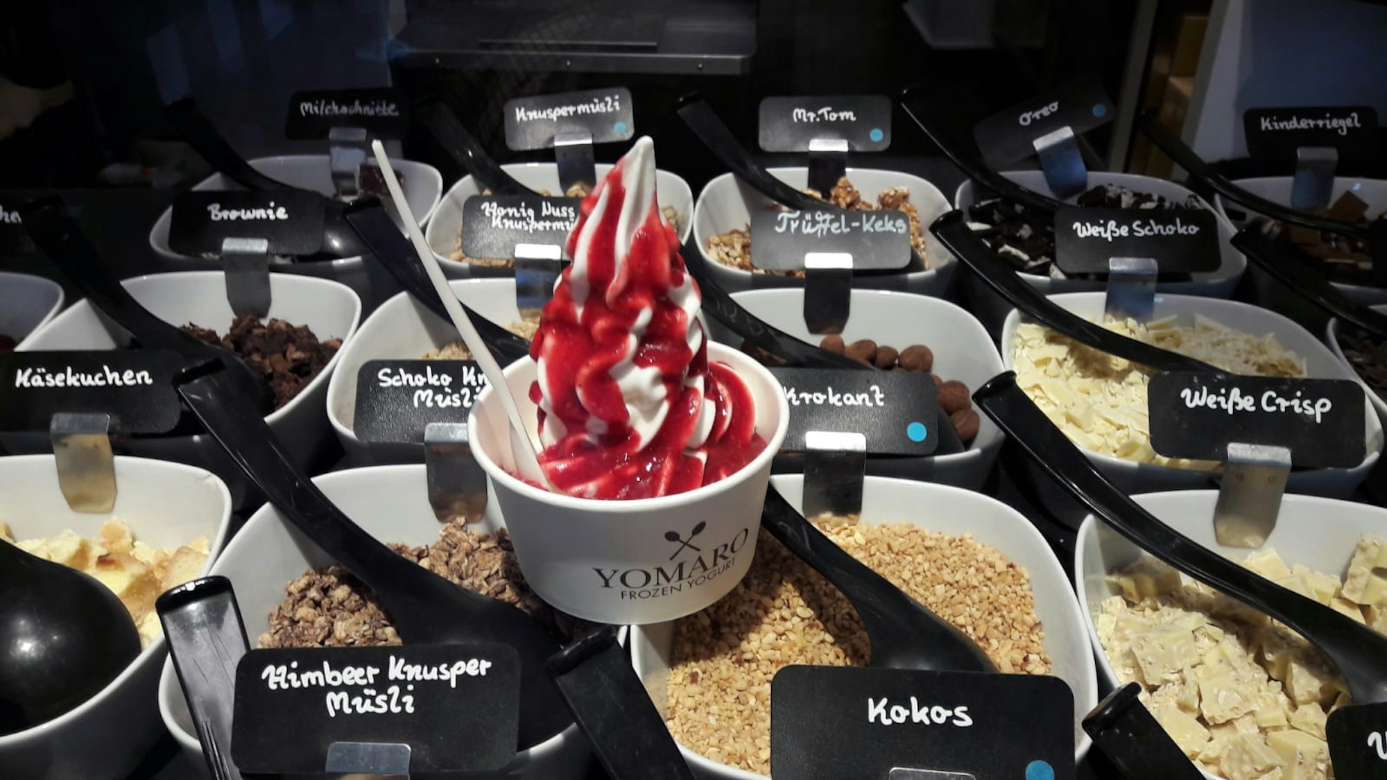 Ein Yomaro-Becher mit Frozen Yogurt und roter Fruchtsoße steht auf der Theke. Darunter sind verschiedenste Toppings zu sehen.