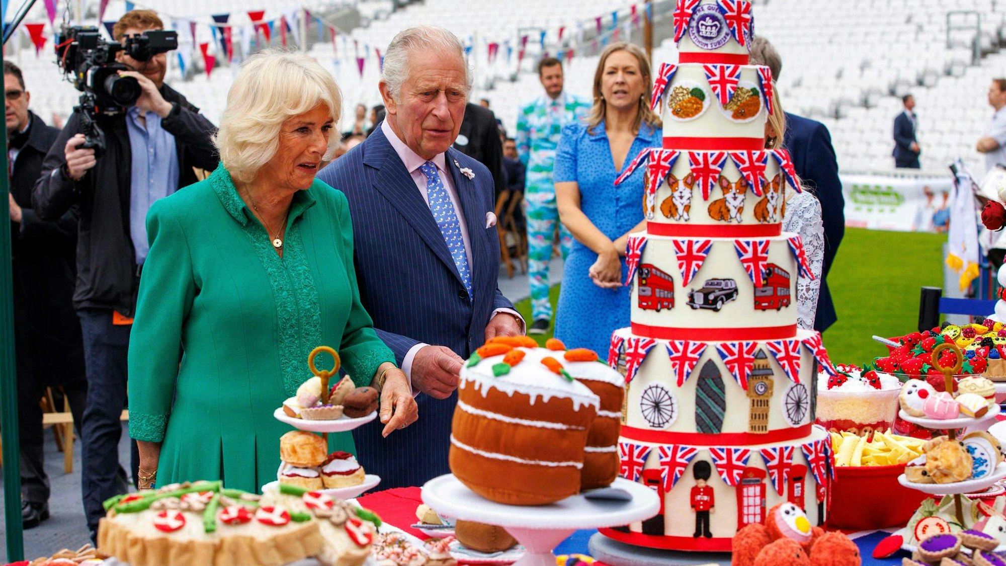 Königin Camilla und König Charles stehen vor einem Tisch mit reich verziertem Gebäck, Kuchen und Torten.