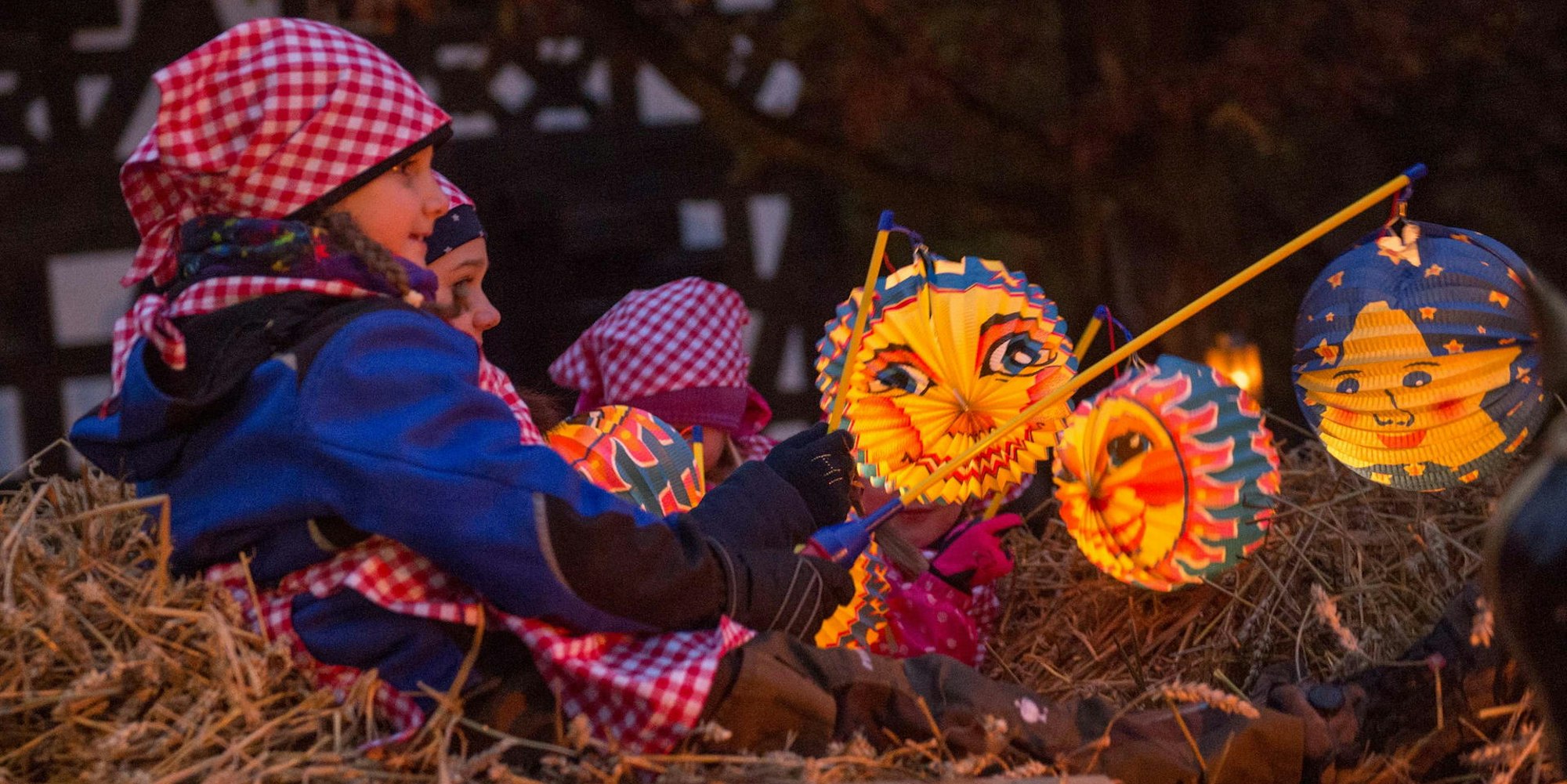 Kinder sitzen im Stroh mit bunten Sonne-, Mond- und Sterne-Laternen.