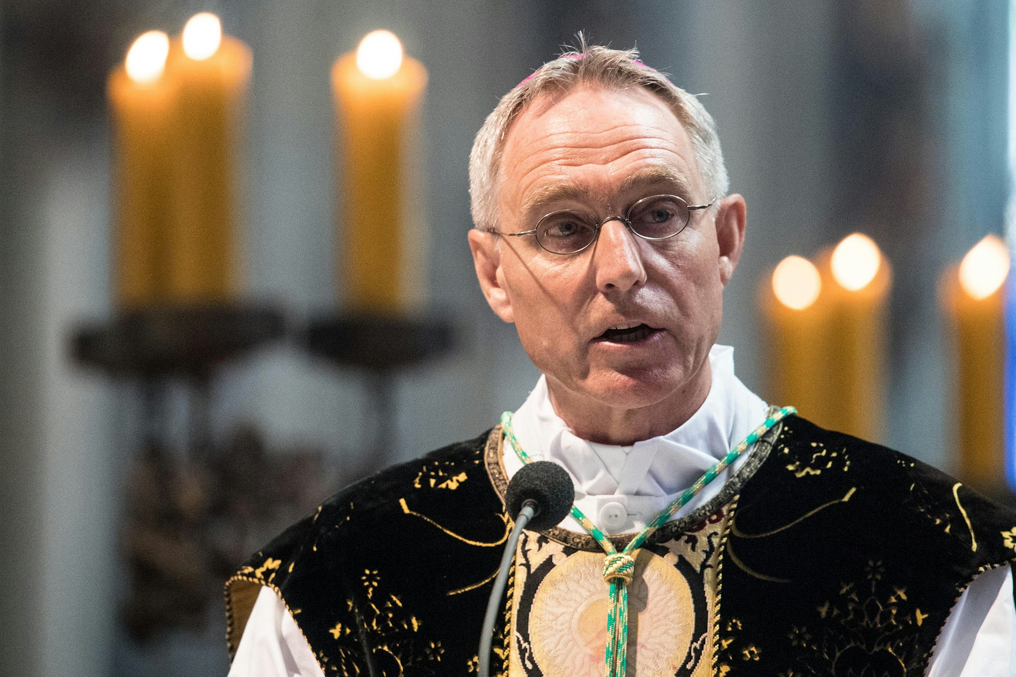 Georg Gänswein in einer schwarzen Casel (barockes Messgewand) mit goldene Besatz vor einem Mikrofon. Im Hintergrund sind unscharf Kerzen zu erkennen.