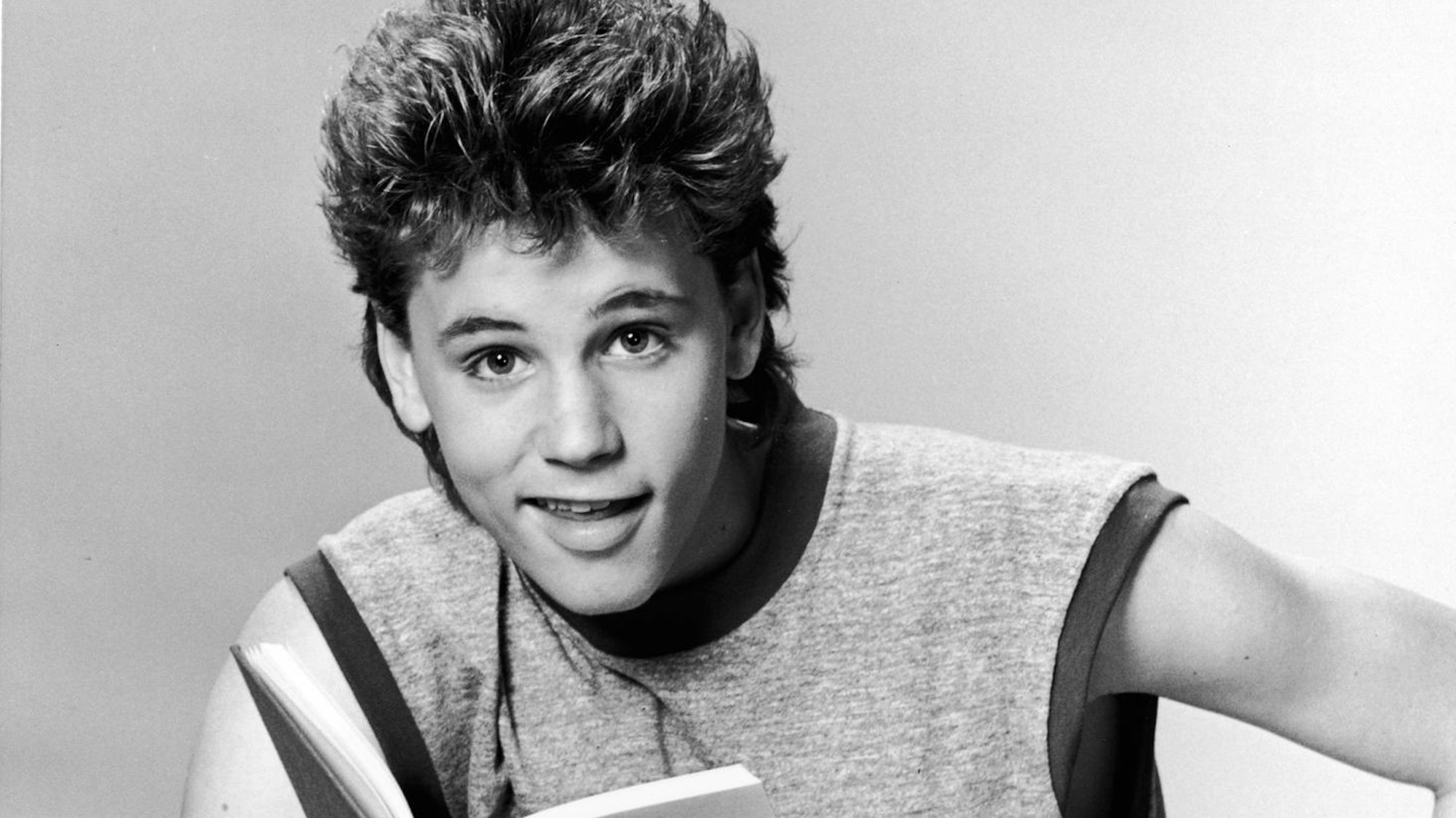 Corey Haim mit Buch. Er trägt ein Sport-Tanktop und schaut lachend in die Kamera.