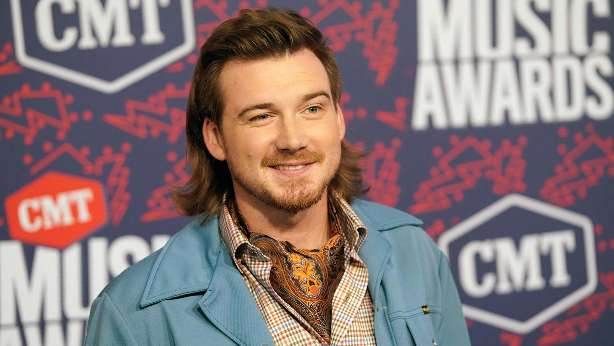 Morgan Wallen mit Vokuhila, babyblauer Stoffjacke und Halstuch. Er grinst smart in die Kamera.