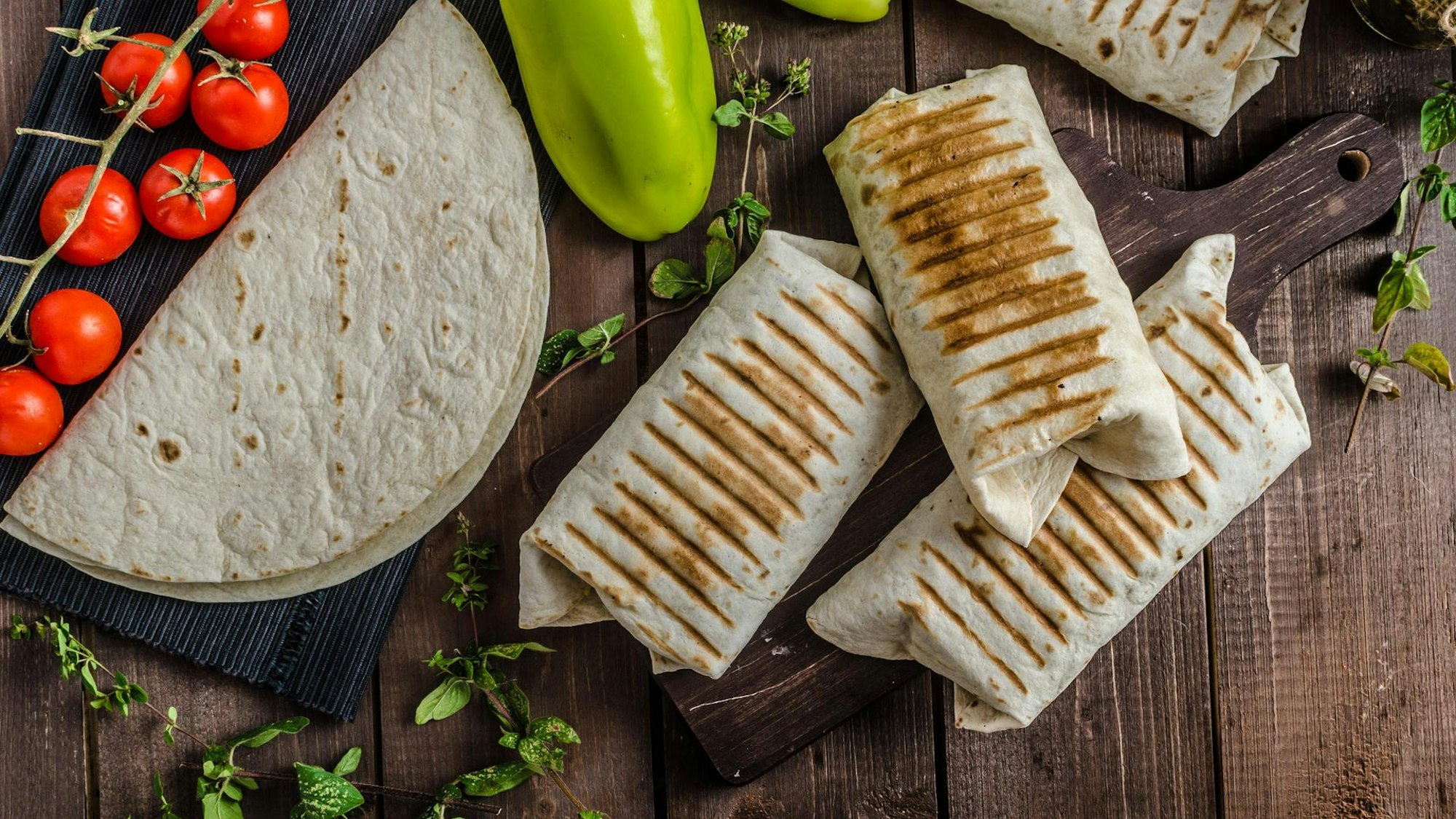 Drei Burritos liegen nebeneinander, daneben ein Tortillafladen und Tomaten.