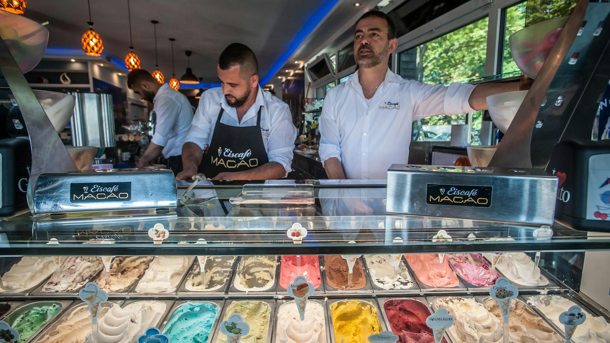 Hinter der Eistheke der Eisdiele Macao arbeiten mehrere Männer, im Vordergrund sind Eissorten zu sehen.