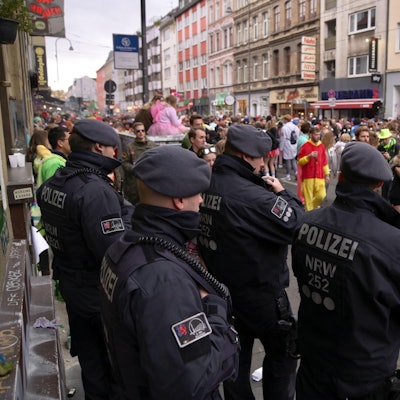 Polizisten einer Hundertschaft stehen am Rand der Zülpicher Straße und beobachten kostümierte Menschen, die vorüberziehen.