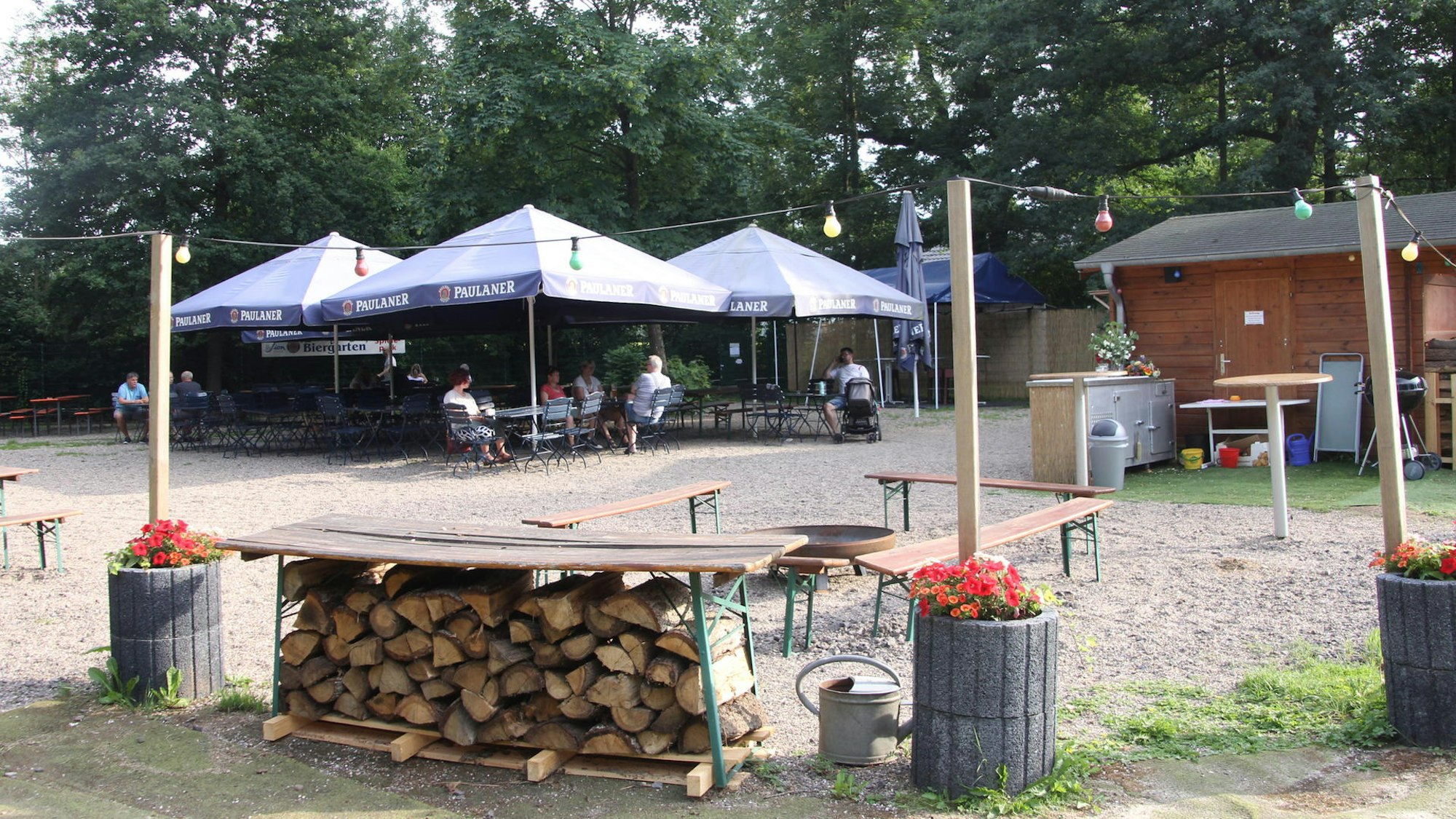 Auf einem Platz stehen Tische und Stühle unter Sonnenschirmen, mehrere Personen sitzen dort. Außenrum führt eine Lichterkette.