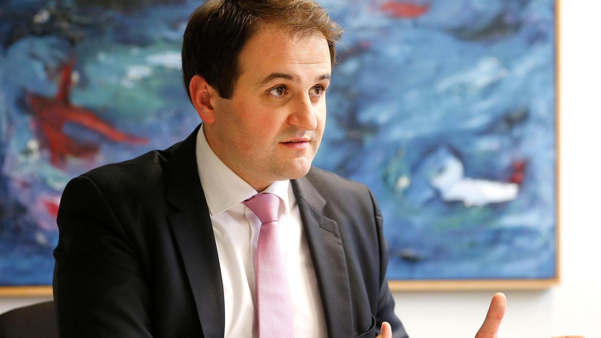 NRW-Europaminister Nathanael Liminski (CDU) in einer Gesprächssituation, den Gesprächspartner sieht man nicht. Er sitzt vor einem großen abstrakten Gemälde mit viel Blau, trägt einen dunklen Anzug, weißes Hemd und rosafarbene Krawatte.
