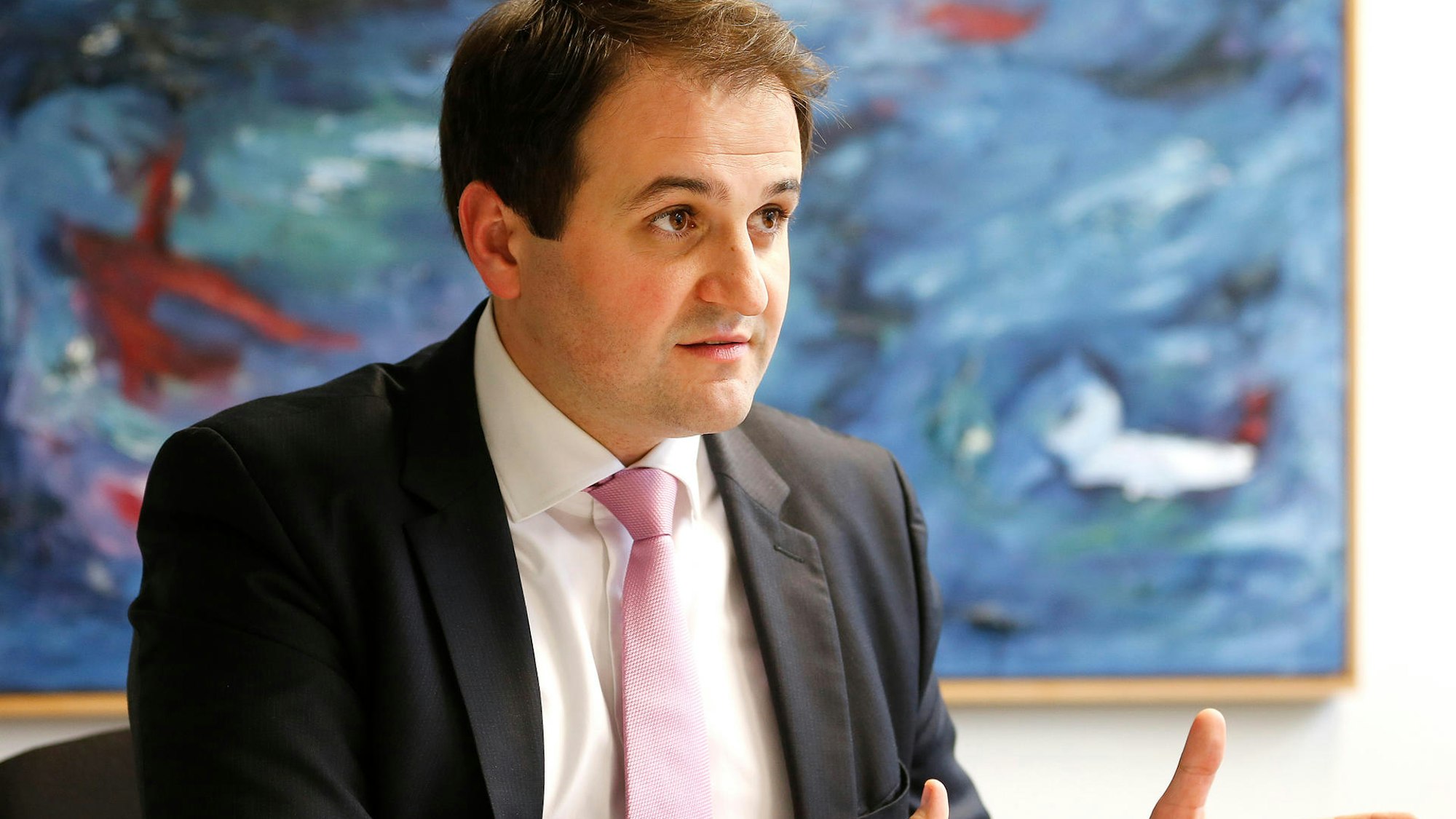 NRW-Europaminister Nathanael Liminski (CDU) in einer Gesprächssituation, den Gesprächspartner sieht man nicht. Er sitzt vor einem großen abstrakten Gemälde mit viel Blau, trägt einen dunklen Anzug, weißes Hemd und rosafarbene Krawatte.