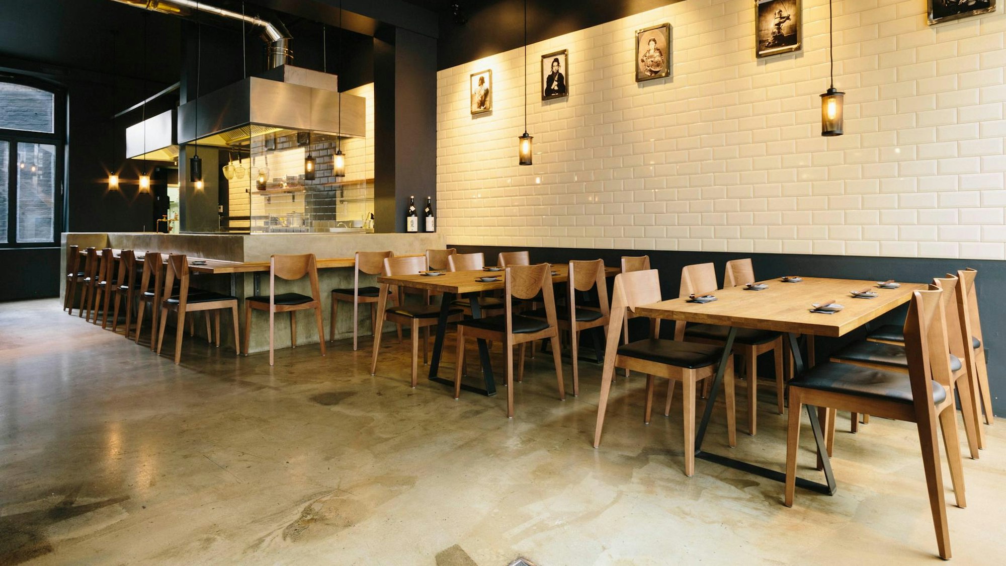 Tische und Stühle aus Holz vor einer weiß gefliesten Wand in einem schlicht gestalteten Raum