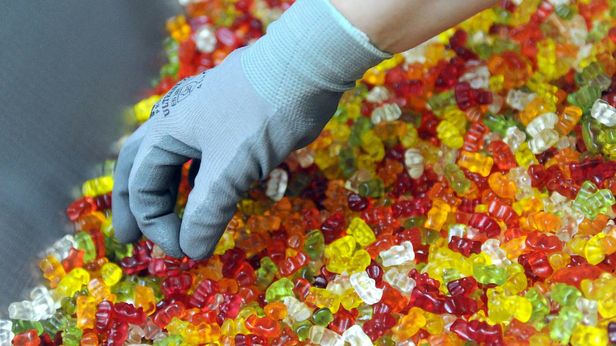 Hand mit Handschuh greift in eine große Menge Gummibärchen