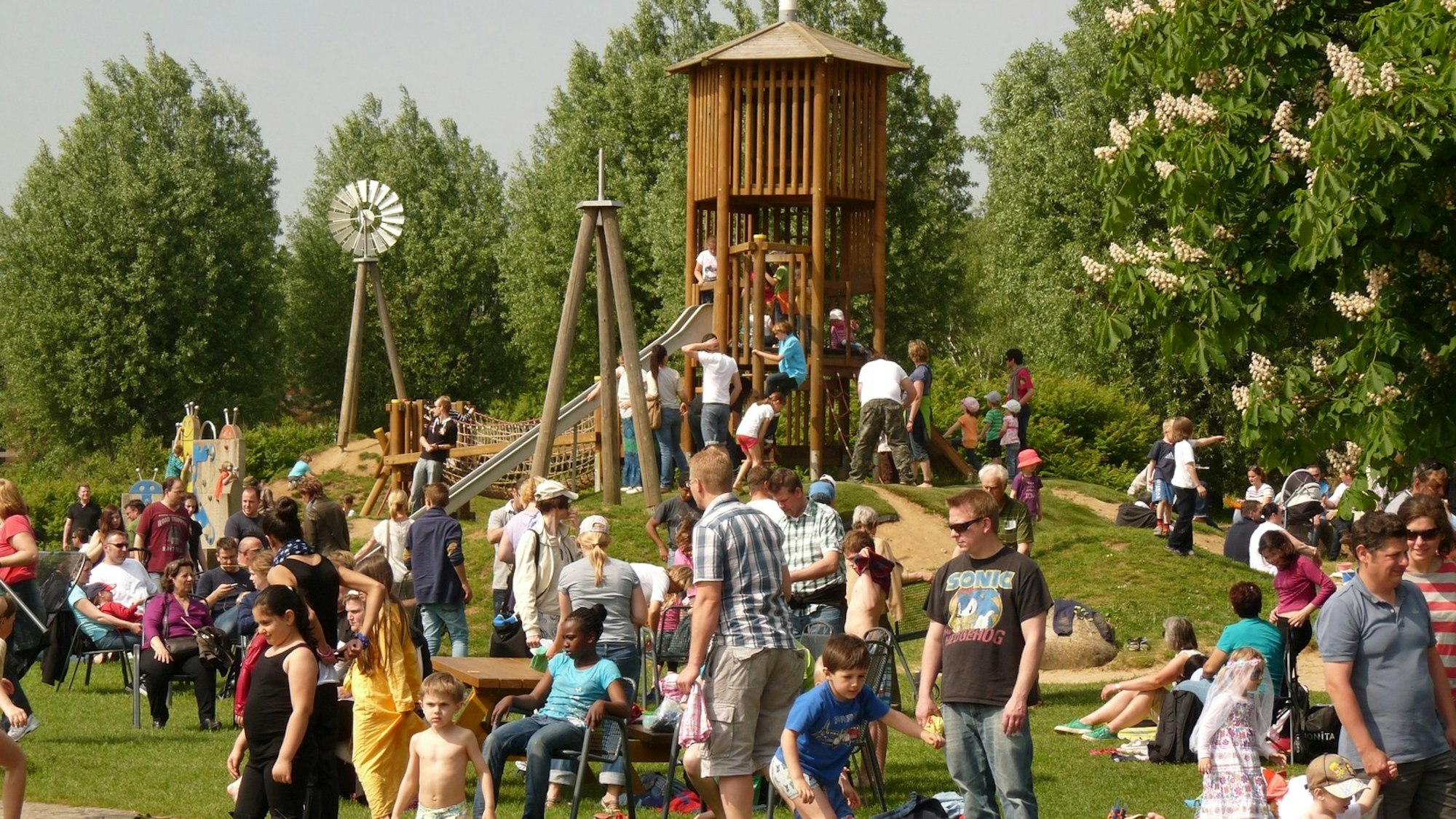 Kinder und Erwachsene auf einer Wiese, im Hintergrund ein Holzturm mit Rutsche.