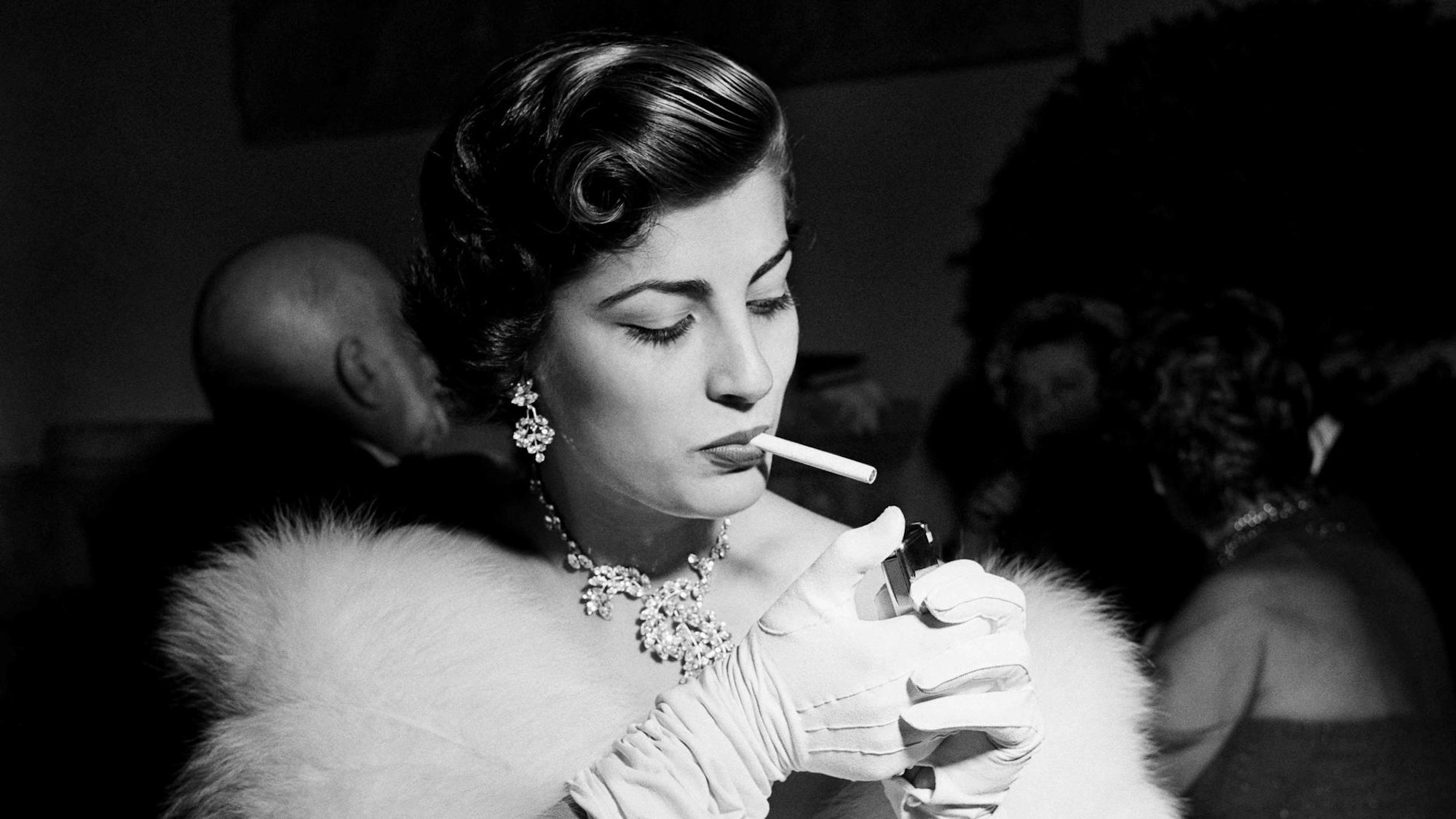 Irene Papas mit Zigarette auf einer Party. Sie trägt Pelz und weiße Handschuhe.
